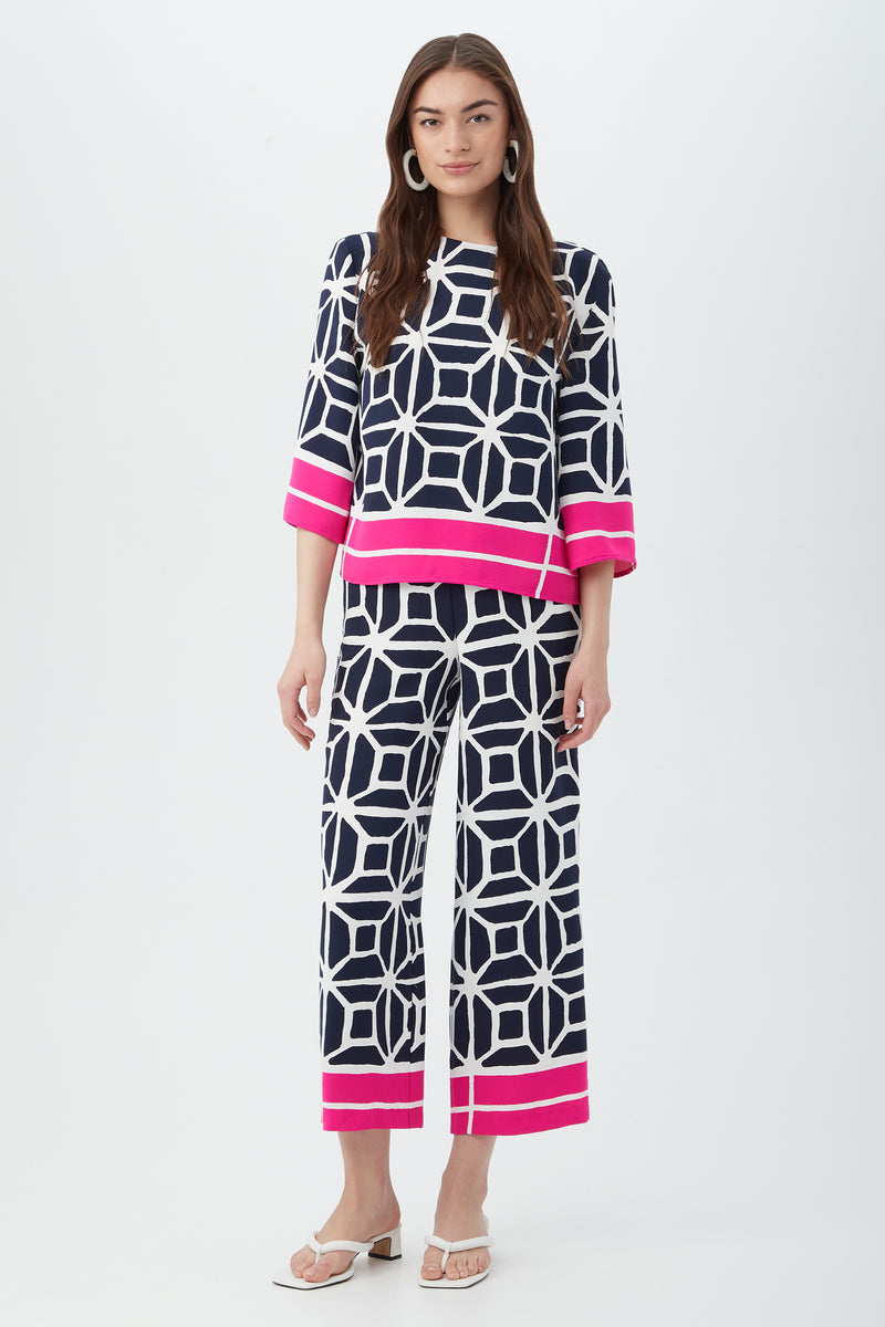 A woman wears the Orlett Wide-Leg Crop Pant in Durell Georgette, paired with a matching navy and white geometric print top featuring bright pink borders. She has loose brown hair, white earrings, and white sandals.