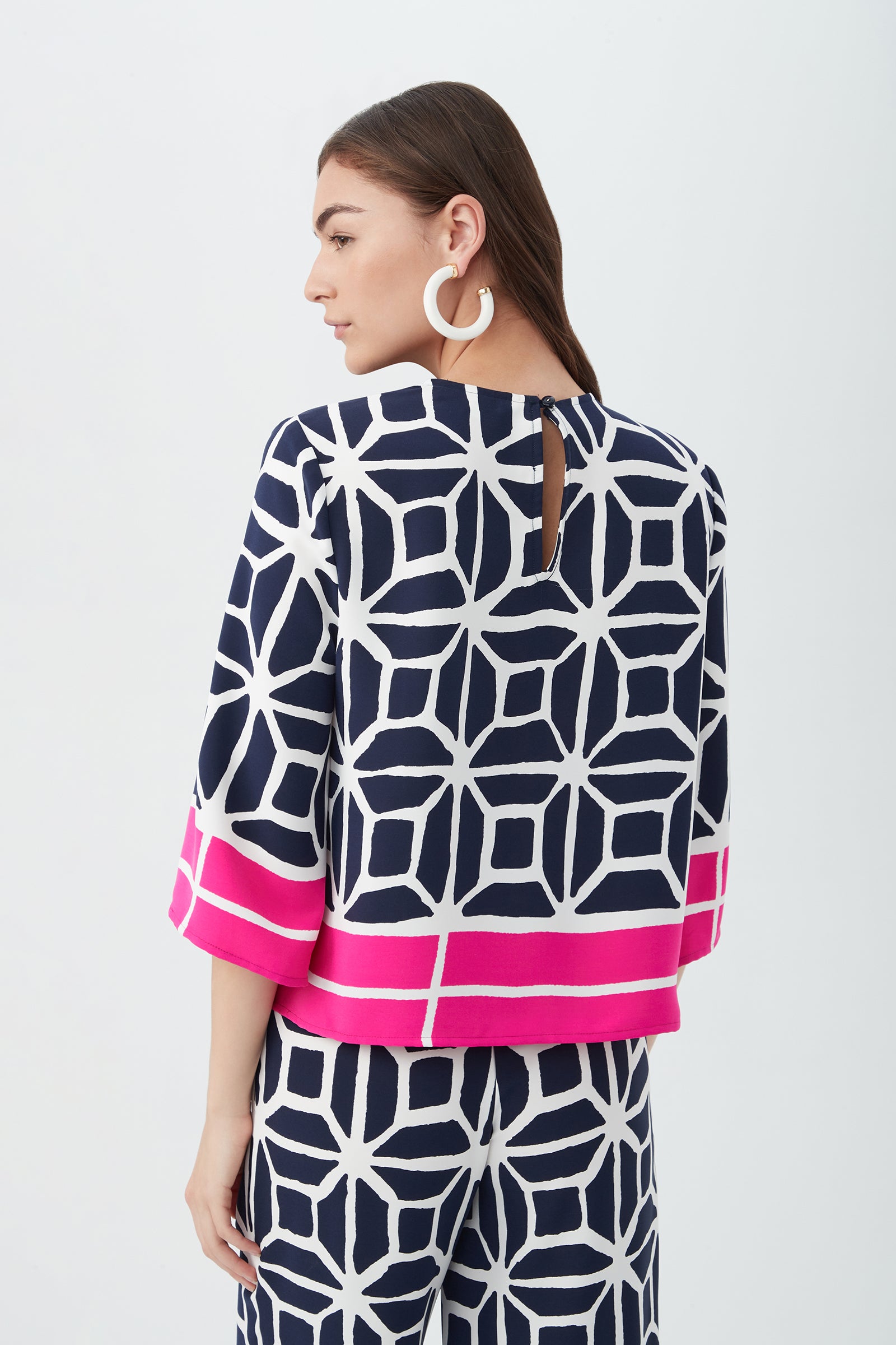 A woman with long brown hair is seen from behind wearing the Muria 3/4-Sleeve Top - Durell Georgette, featuring a navy and white geometric pattern with wide pink stripes at the cuffs and hem, paired with matching pants and large circular earrings.