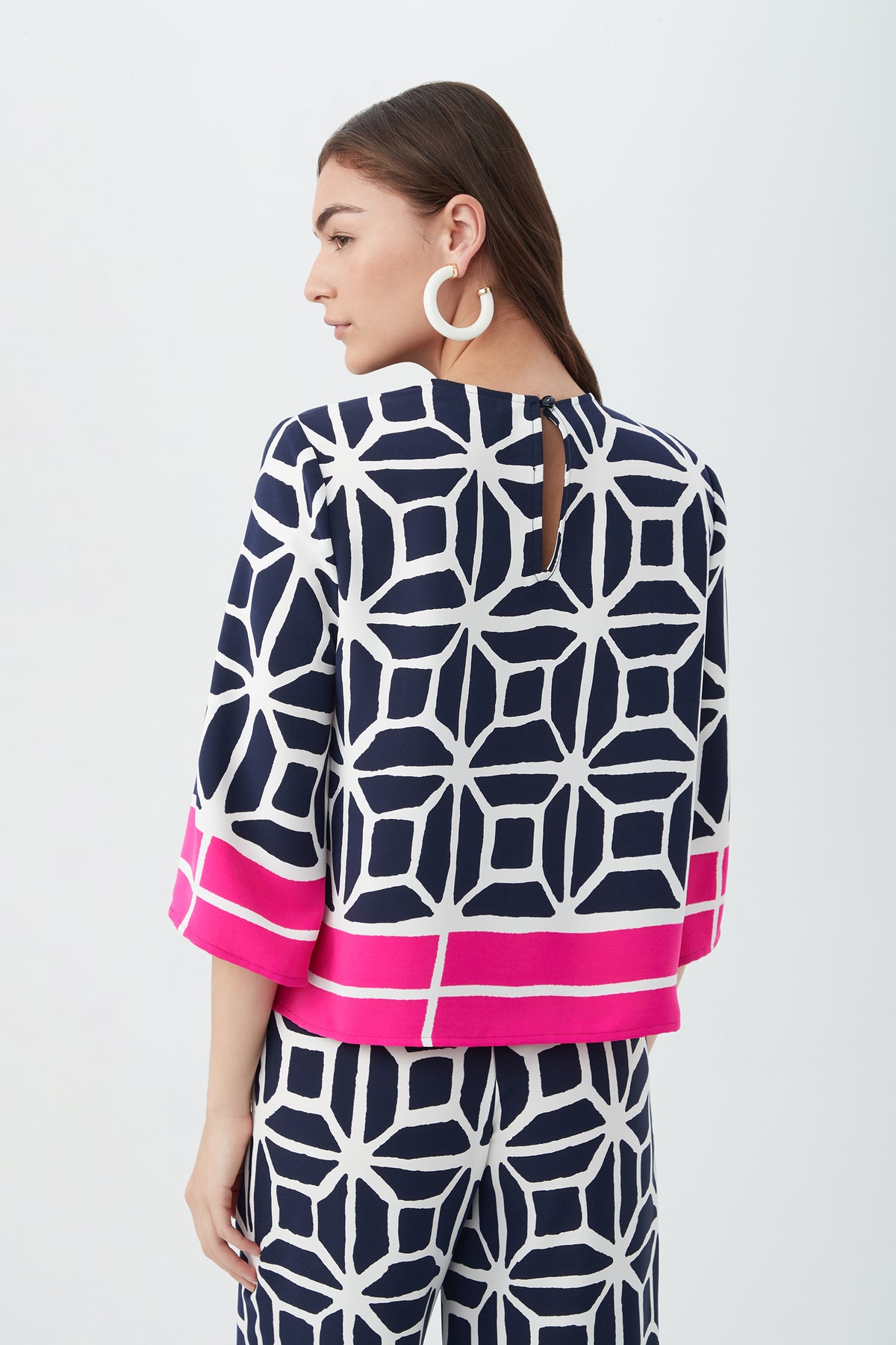 A woman with long brown hair is seen from behind wearing the Muria 3/4-Sleeve Top - Durell Georgette, featuring a navy and white geometric pattern with wide pink stripes at the cuffs and hem, paired with matching pants and large circular earrings.