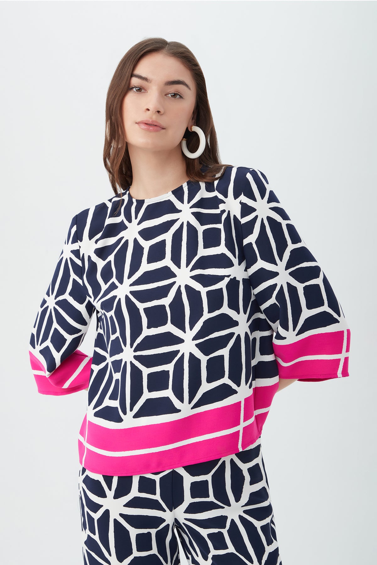 A woman with long brown hair wears the Muria 3/4-Sleeve Top - Durell Georgette in multi, featuring a navy and white geometric print with pink accents, paired with matching pants. She stands against a plain light background, wearing white hoop earrings.