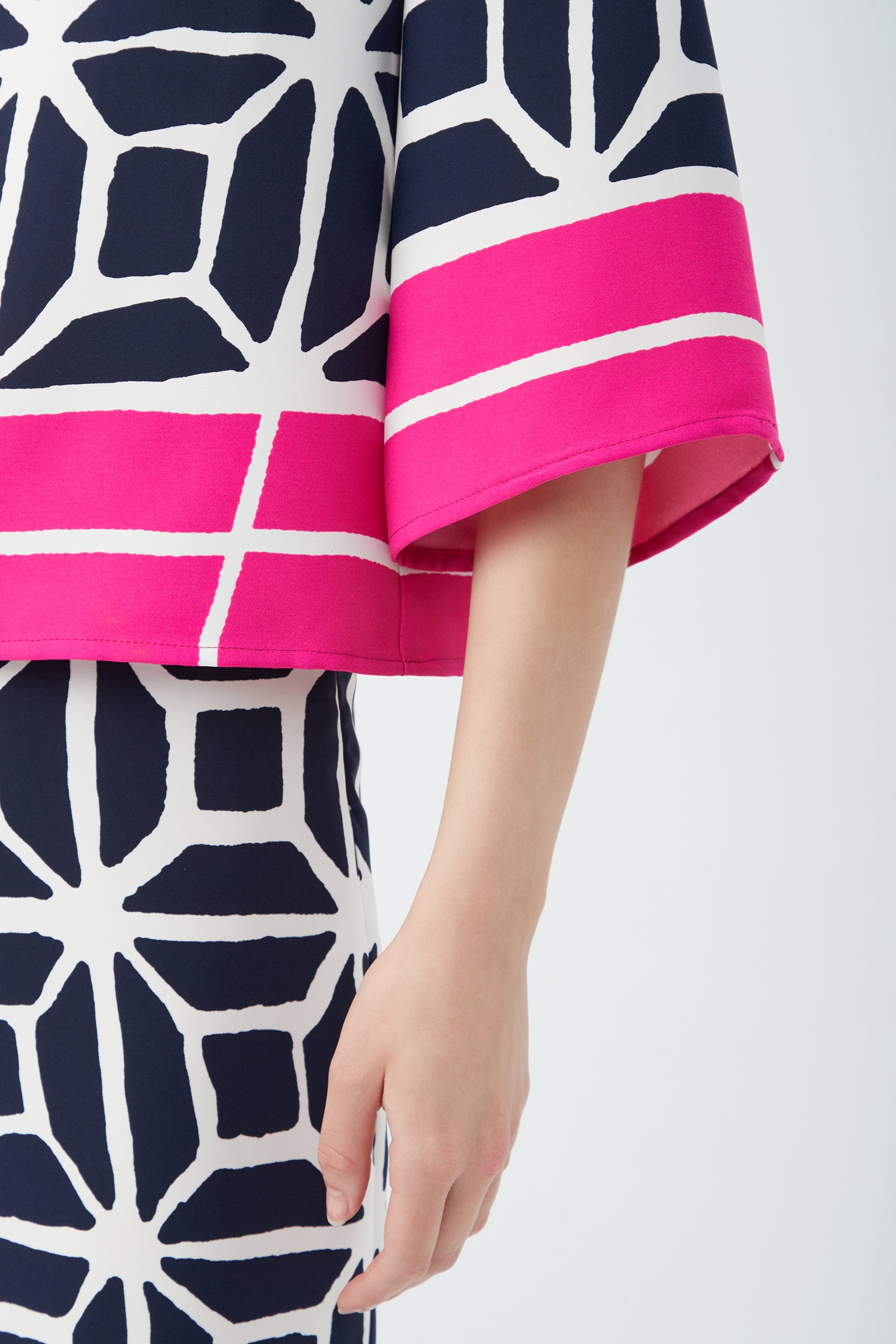 A person wears the Muria 3/4-Sleeve Top - Durell Georgette, featuring a geometric navy and white pattern with bold pink stripes on the top and sleeve. Only the torso and one arm are visible against a plain background.