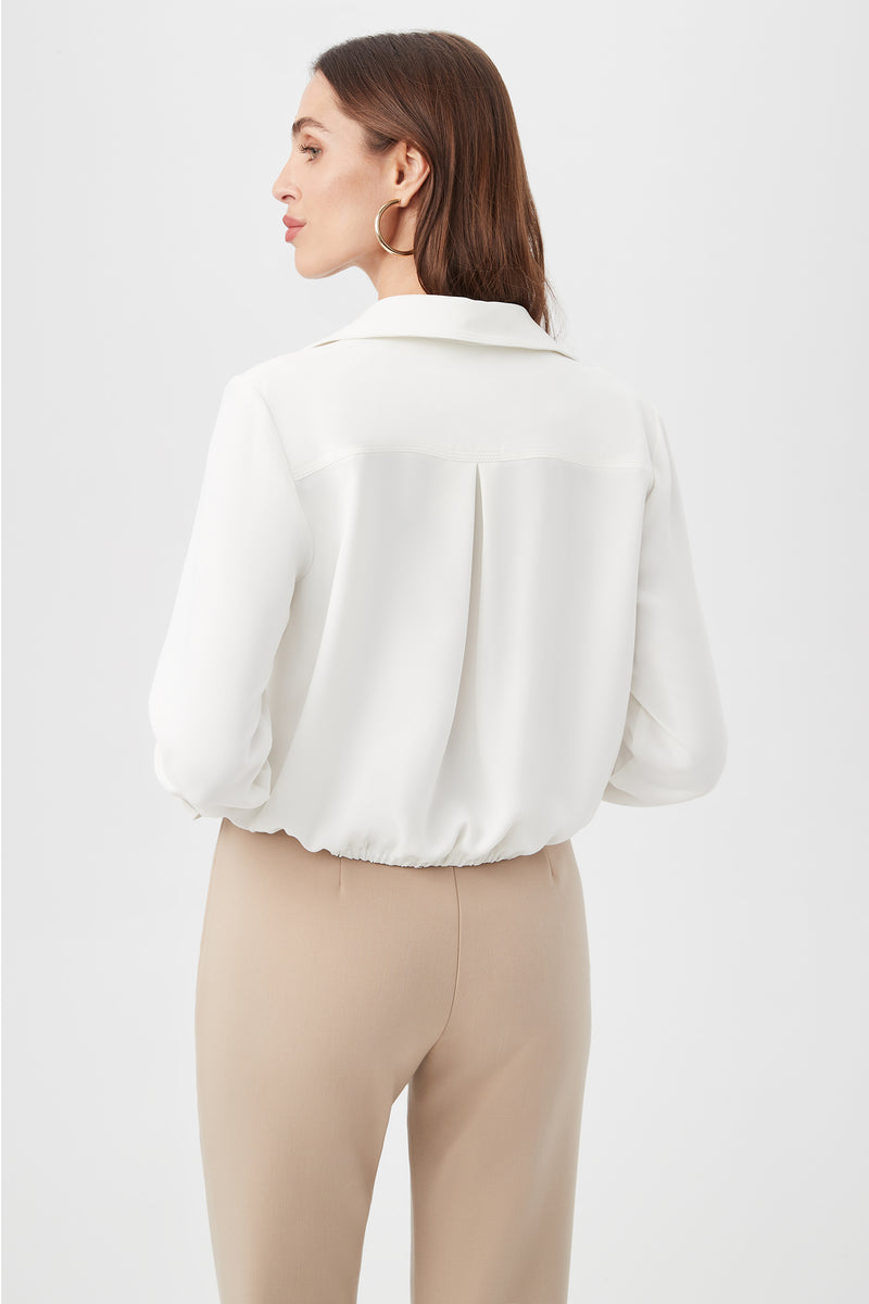A woman with straight brown hair stands with her back to the camera against a white background, wearing the Renlee Shirt, a white long-sleeve blouse, paired with beige pants.