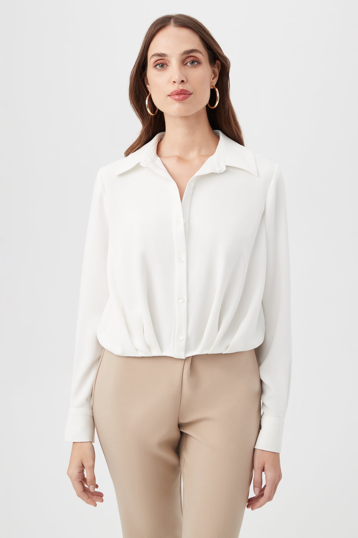 A woman with long brown hair wears gold hoop earrings, beige pants, and the Renlee Shirt—a white collared button-up—while standing against a plain white background and looking at the camera.