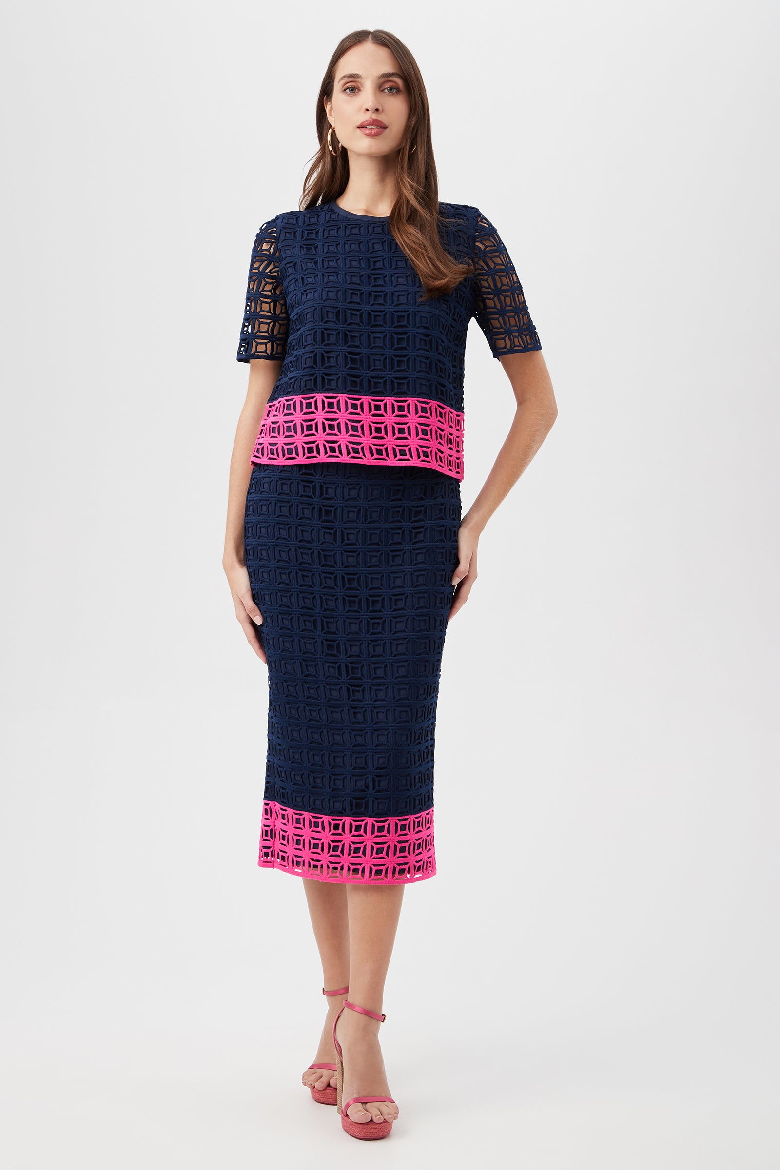 A woman wears the Serilda 2 Top - Block Party Lace, a navy blue lace two-piece with short sleeves, a pencil skirt, and bold pink geometric trim. She poses against a white background with long brown hair and pink sandals.