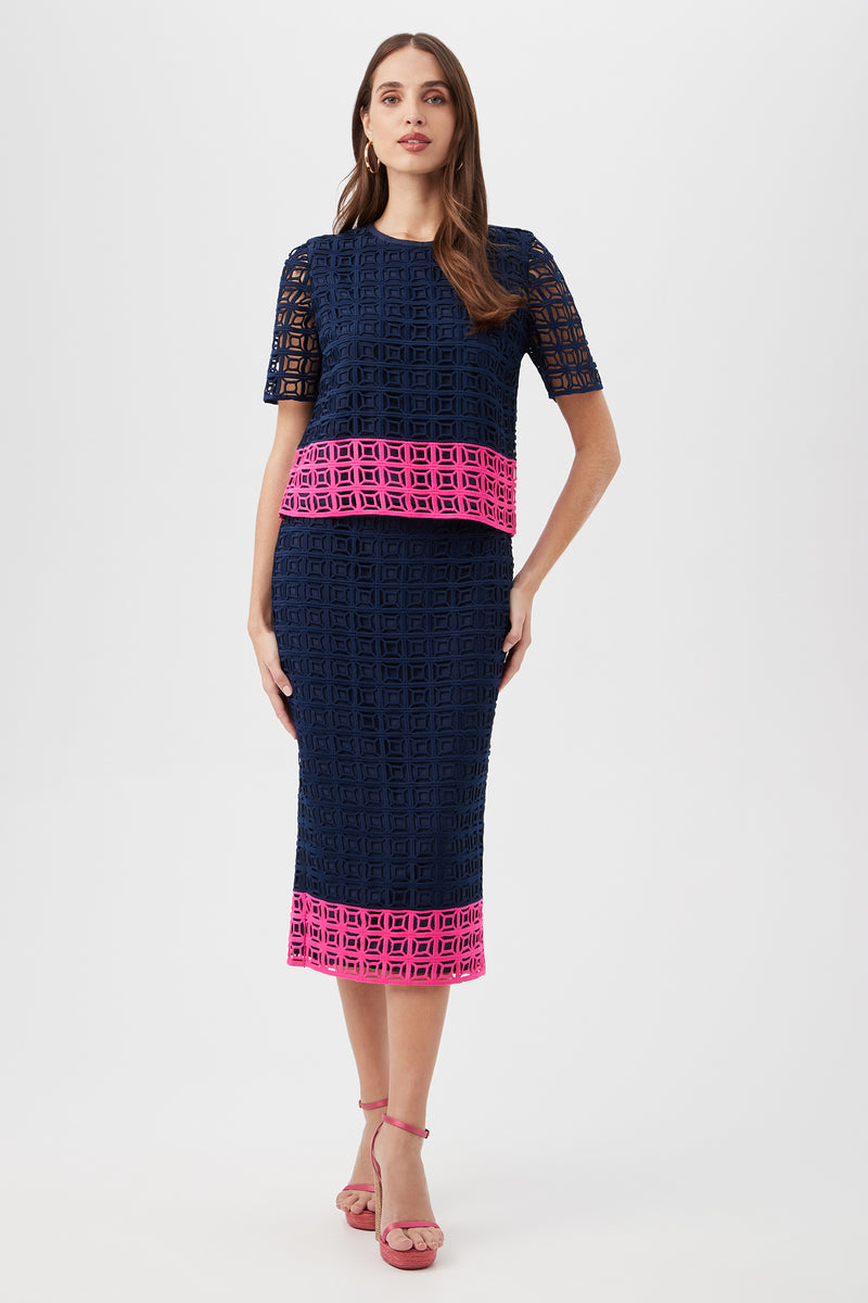 A woman wears the Serilda 2 Top - Block Party Lace, a navy blue lace two-piece with short sleeves, a pencil skirt, and bold pink geometric trim. She poses against a white background with long brown hair and pink sandals.
