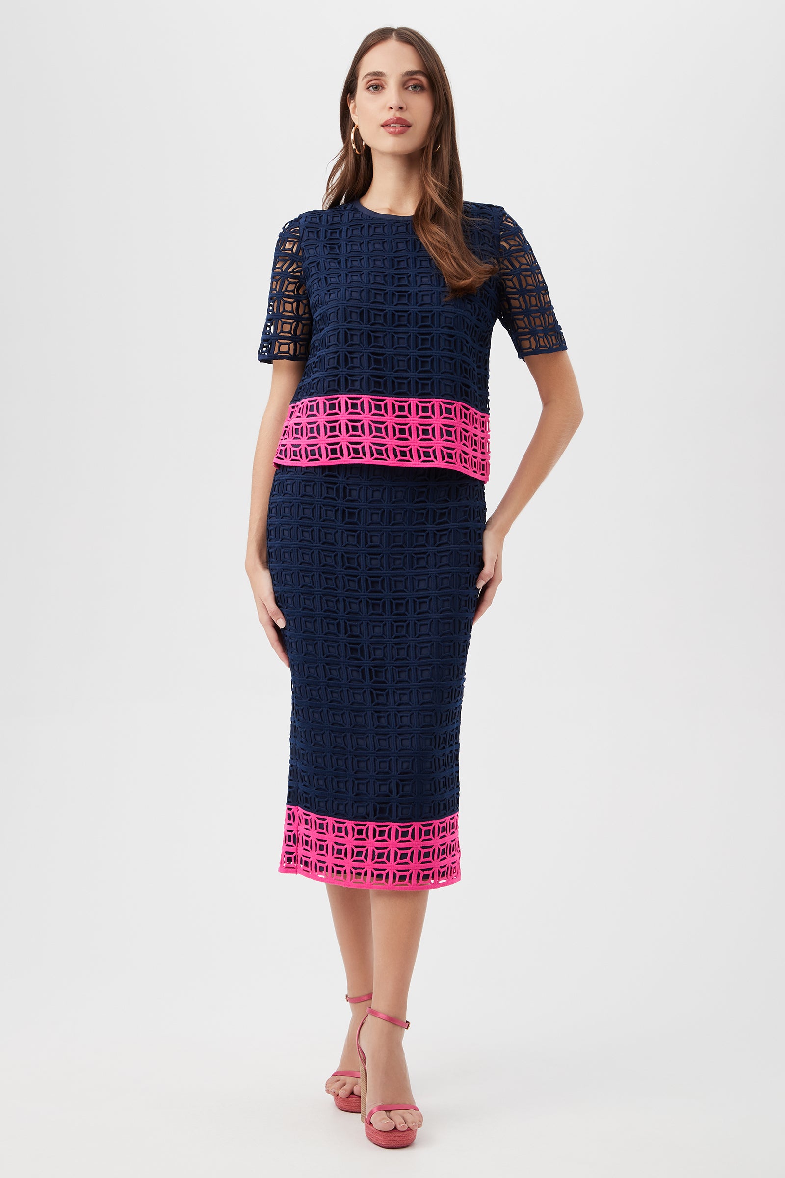 Woman wears the Dita 2 Midi Pencil Skirt - Block Party Lace, featuring a navy geometric pattern with bright pink hem and waist accents, paired with short sleeves and pink strappy heels against a plain background.