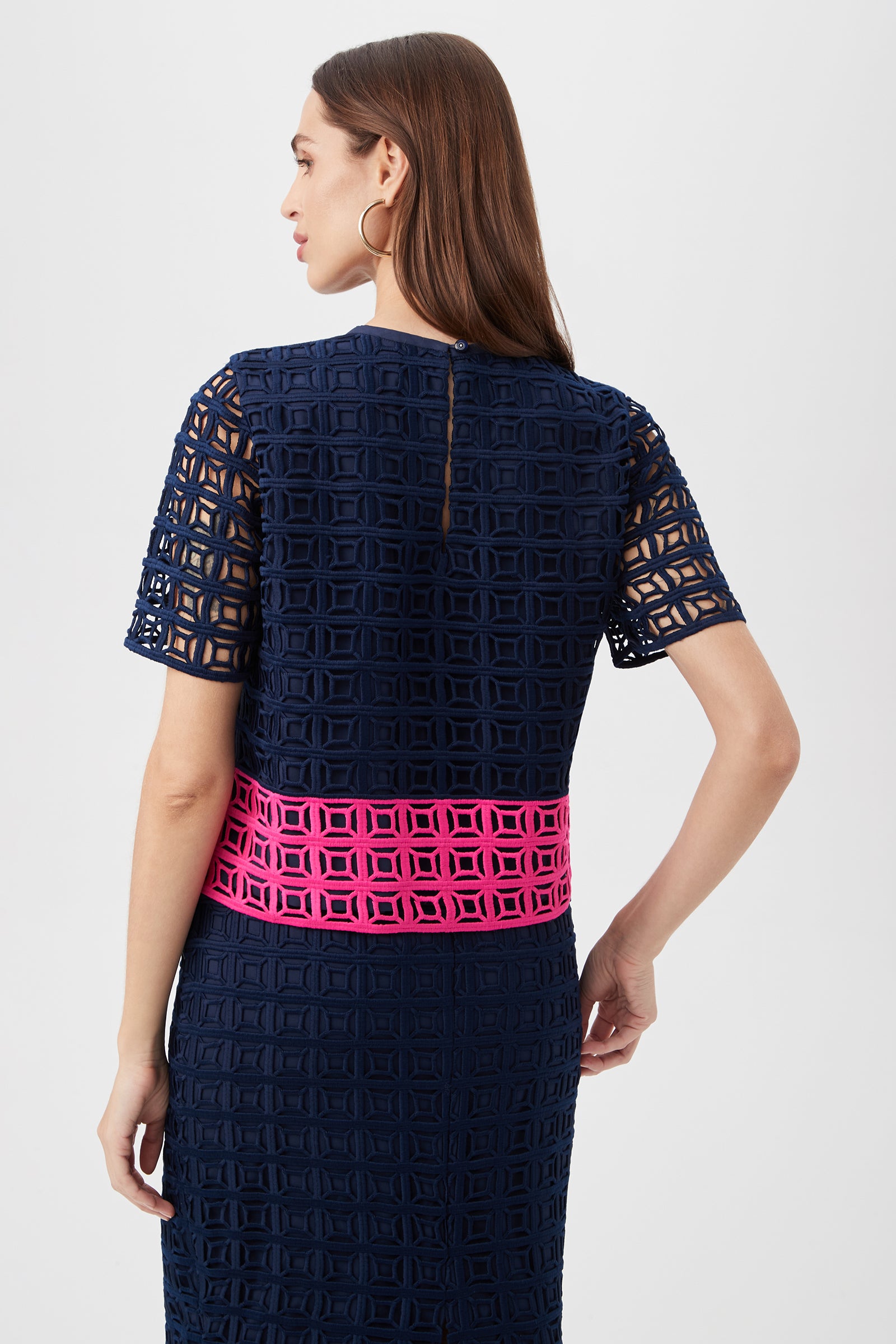 A woman with long brown hair models the Serilda 2 Top – Block Party Lace, featuring a geometric cutout pattern and bright pink waistband, shown from the back against a plain light background.