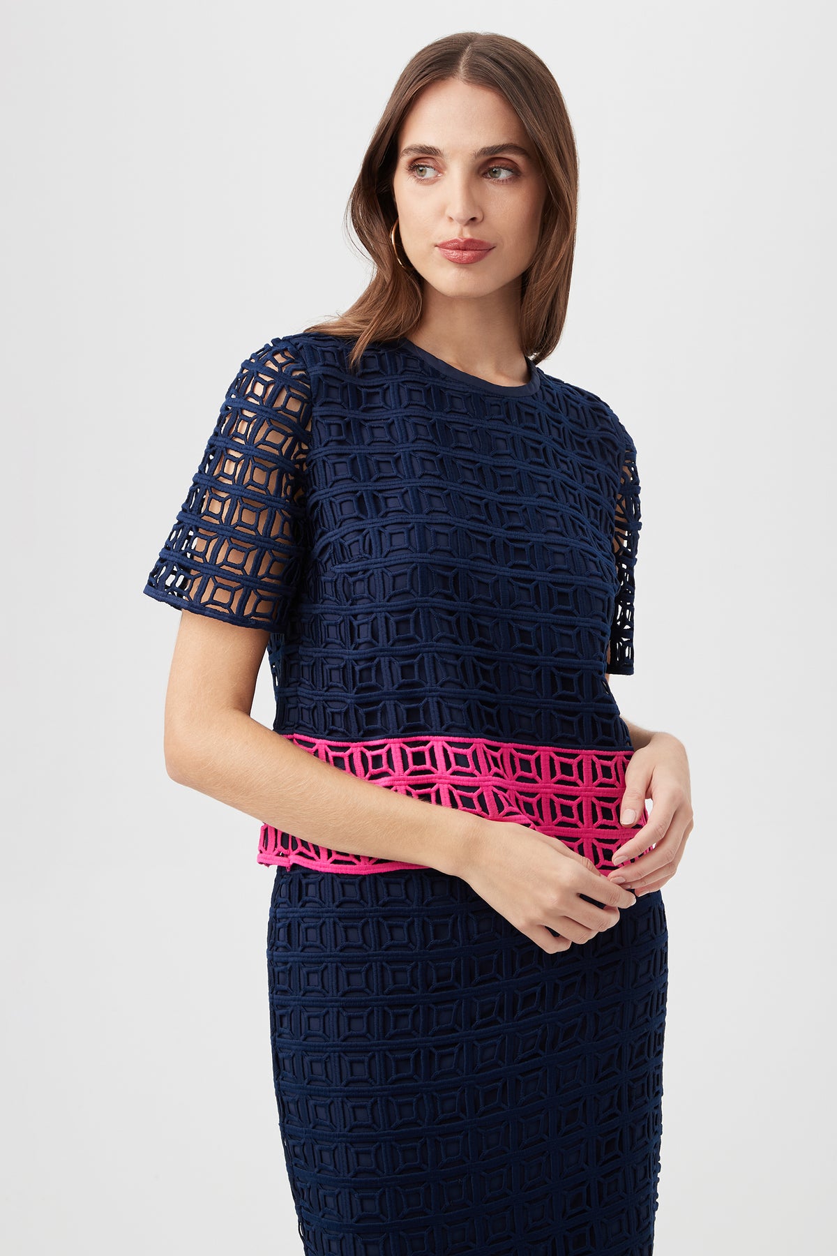 Woman wearing the Serilda 2 Top – Block Party Lace in Indigo/Petal Pink, featuring short sleeves, a geometric cutout pattern, and a bright pink waist band, posed against a plain light background.