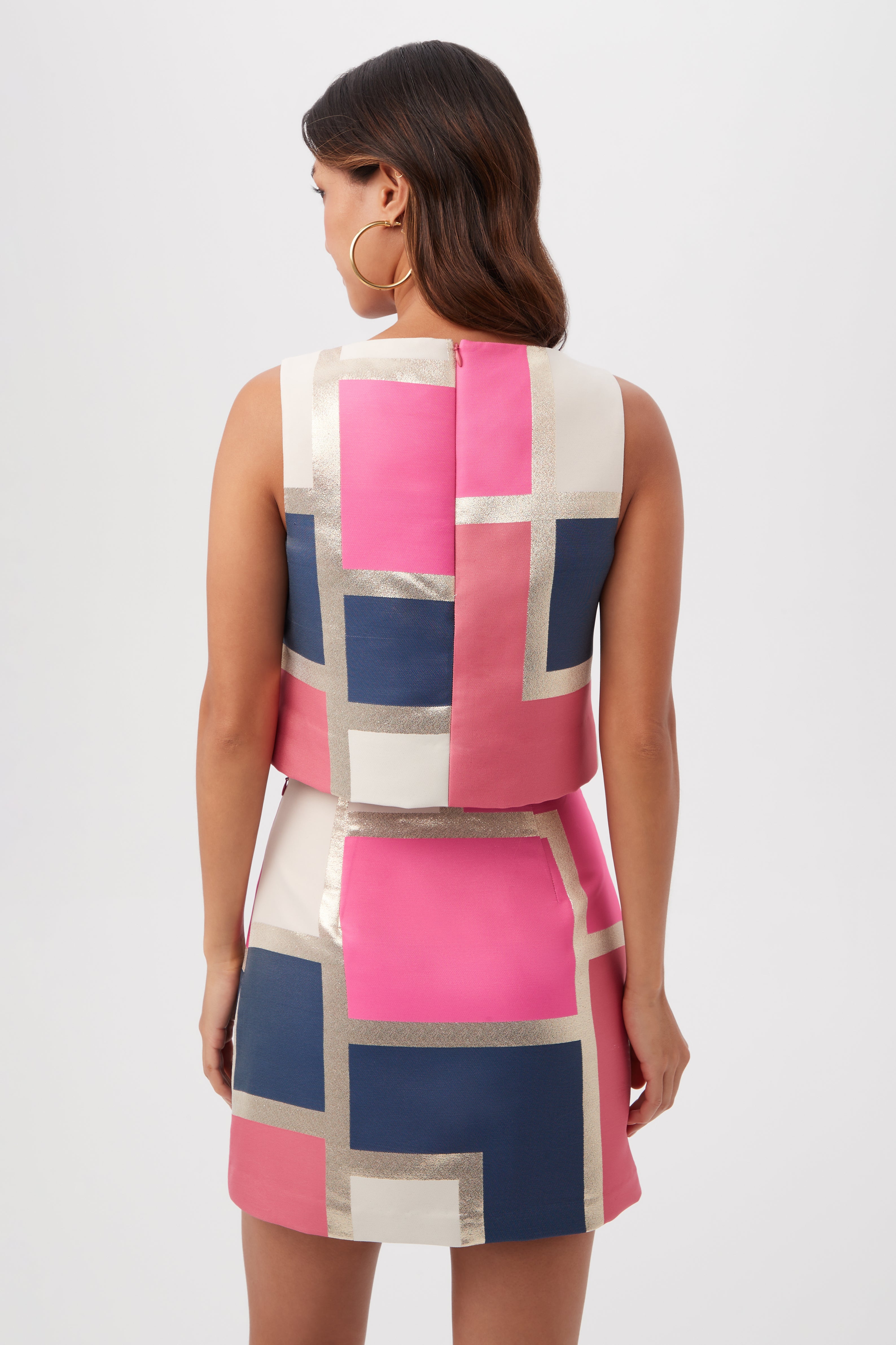 A woman with wavy brown hair faces away, wearing the Cayley 2 Top - Block Party Jacquard, a sleeveless top featuring bold geometric blocks in pink, navy, white, and metallic gold. She accessorizes with large hoop earrings and her arms rest by her sides.