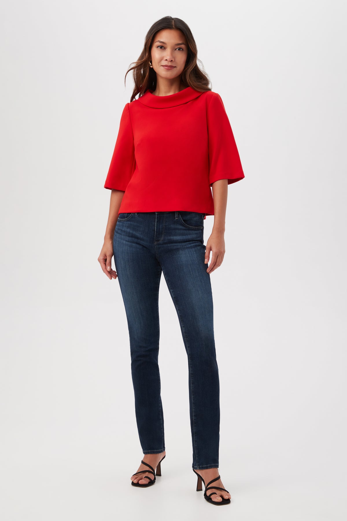 A woman stands against a plain light background, wearing the Kailee Short-Sleeve Folded Collar Top in red, dark blue skinny jeans, and black heeled sandals. She has long wavy hair and looks straight ahead with a slight smile.
