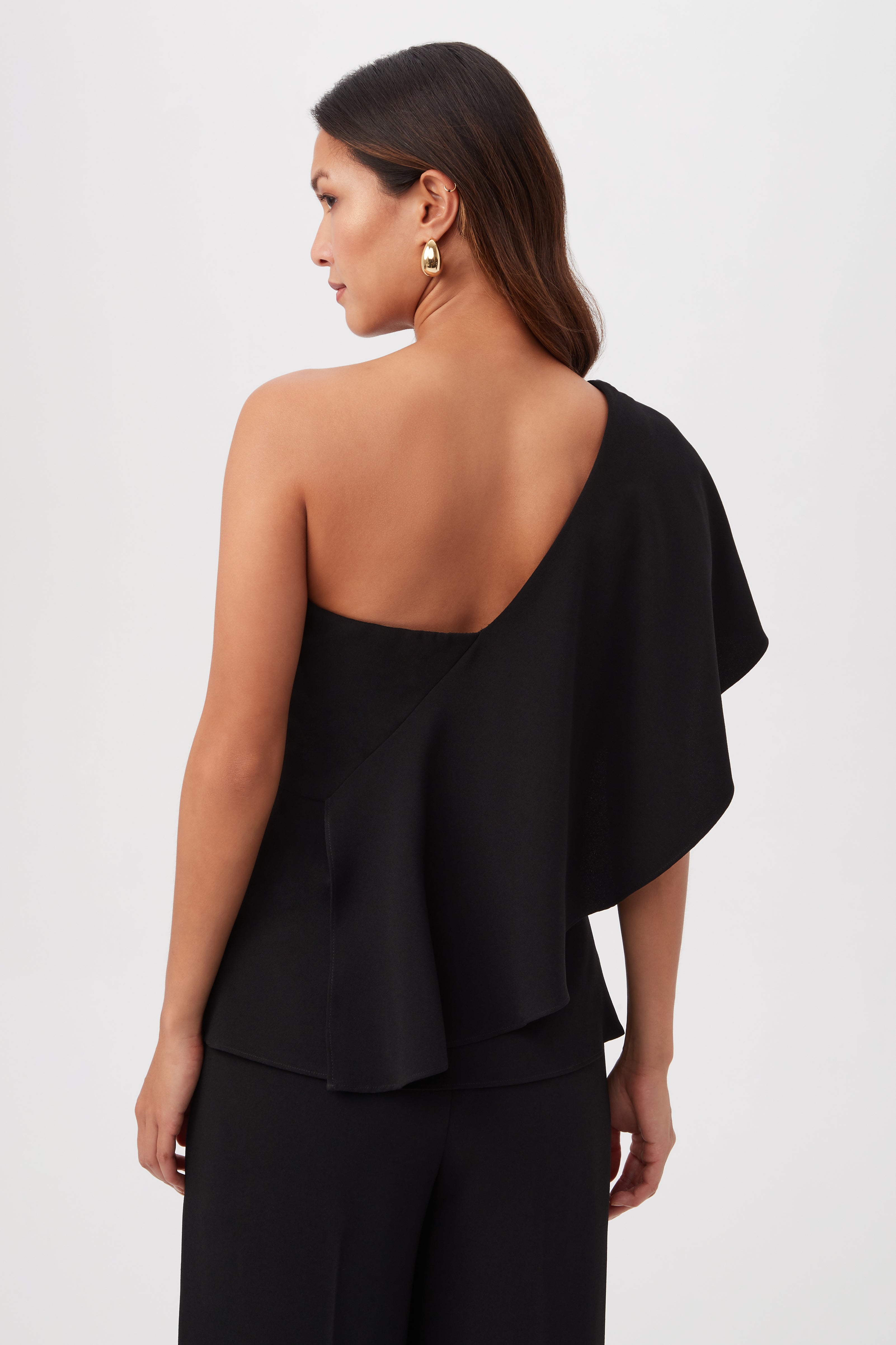 A woman with long brown hair models the Glamorous One-Shoulder Flounce Top and matching black pants, shown from the back against a light background. She completes her look with gold hoop earrings.