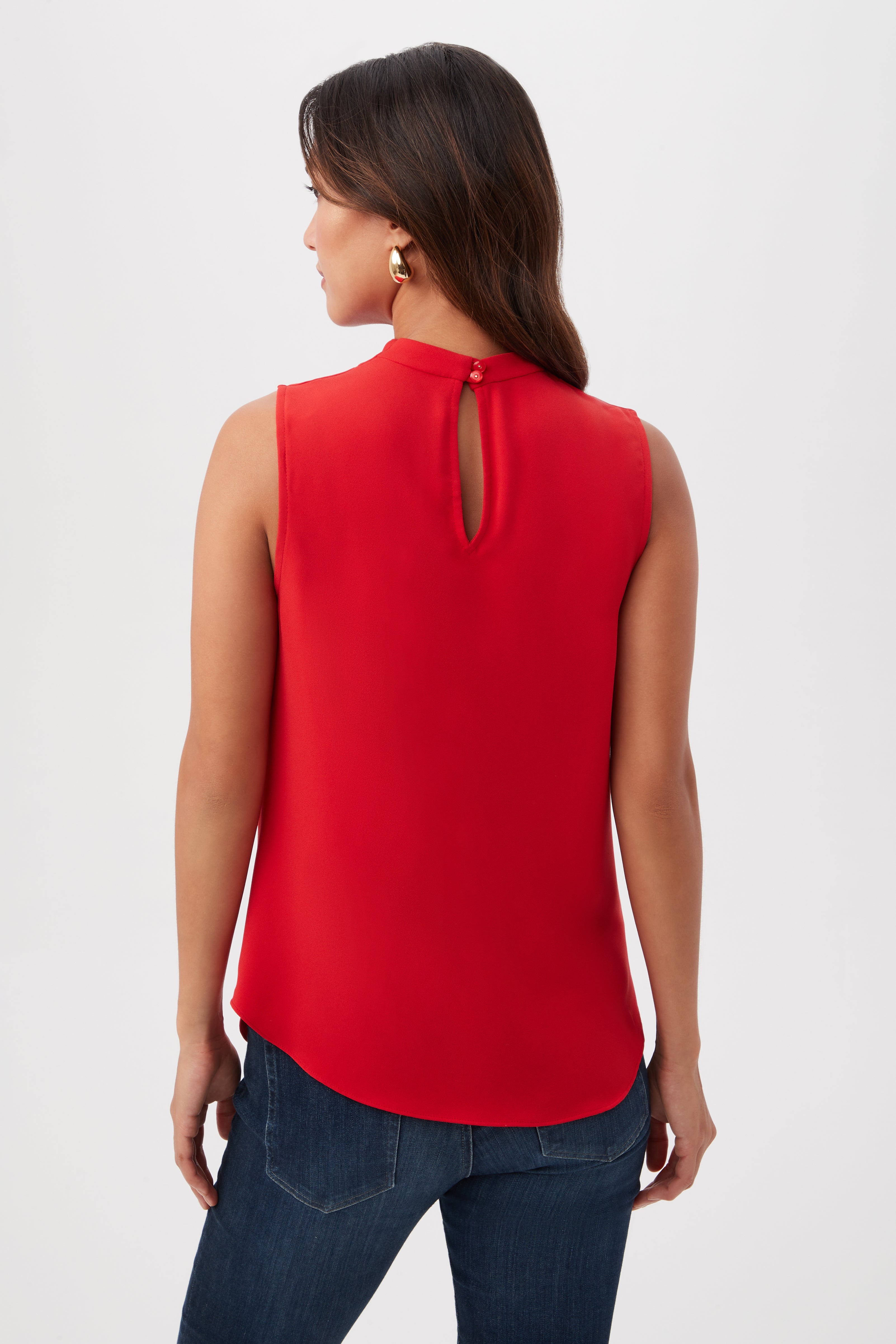 A woman with long dark hair is seen from behind, wearing the Lenaya Sleeveless Cowl Neck Top in red, paired with dark blue jeans. She faces away and looks slightly to the left.