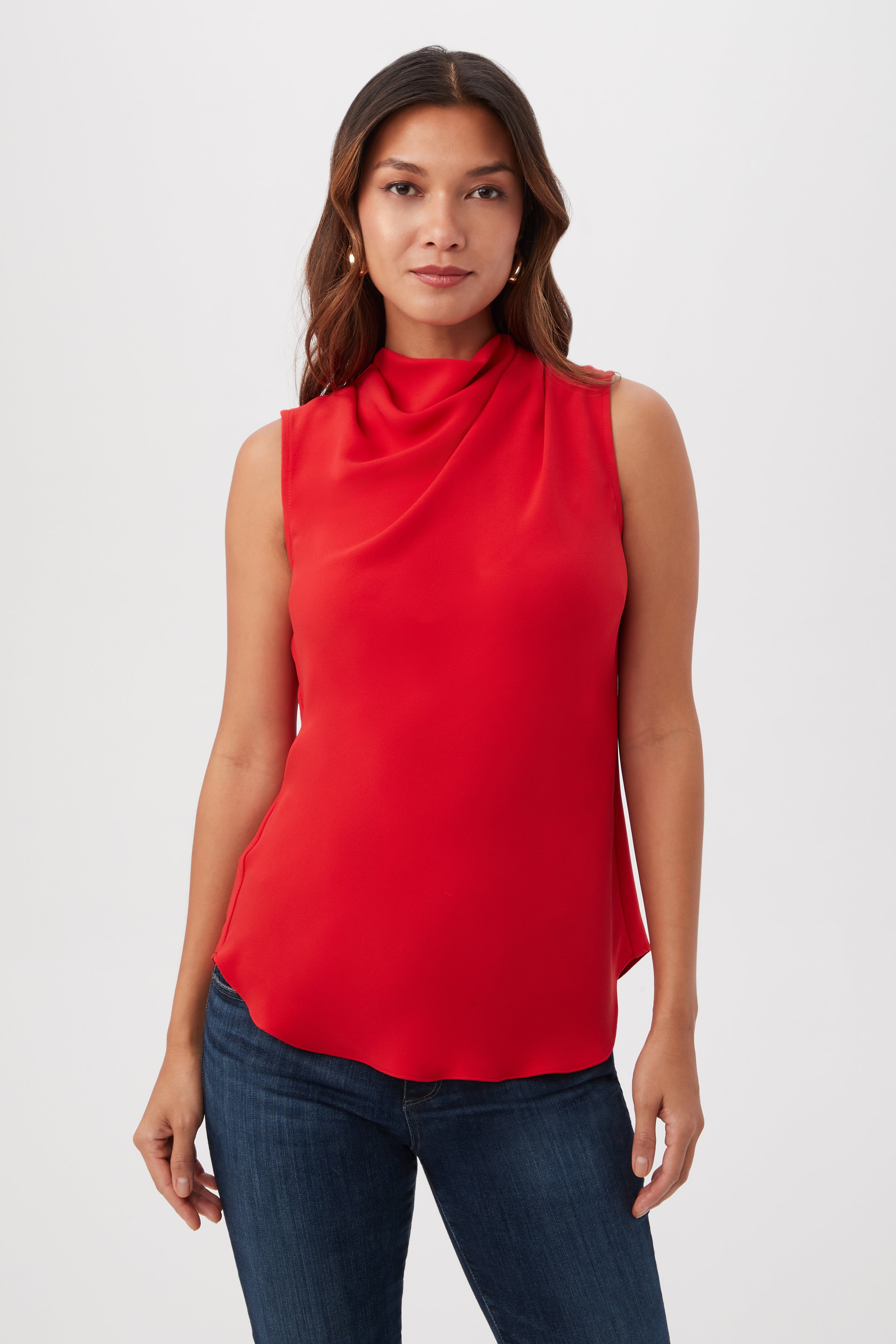 A woman with long brown hair models the Lenaya Sleeveless Cowl Neck Top in Reina Red, paired with blue jeans, standing against a plain white background.