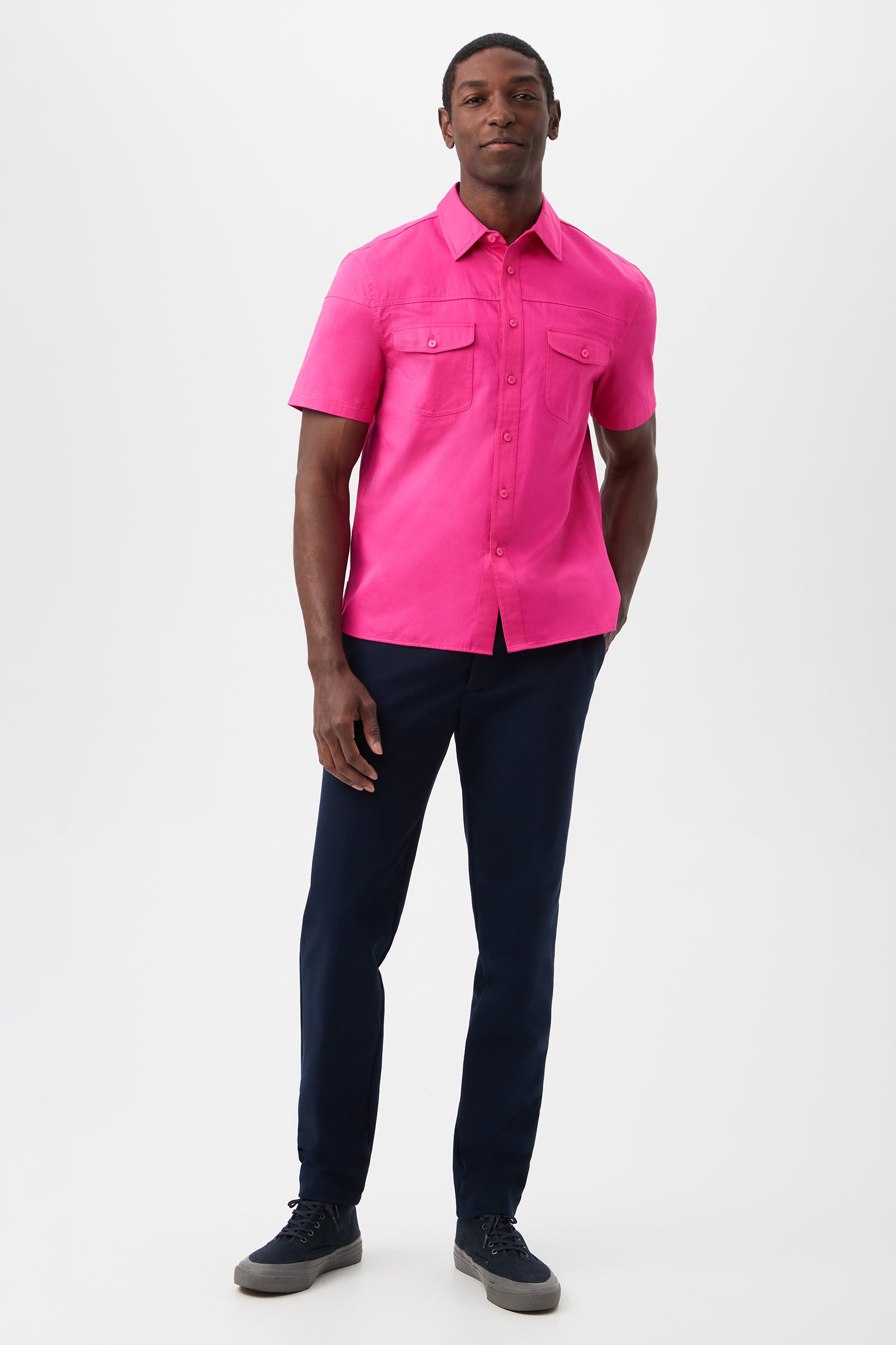 A man stands against a white background wearing the Shelton Short-Sleeve Shirt - Cotton Sateen in bright pink, paired with dark navy pants and sneakers, one hand in his pocket and a relaxed expression.