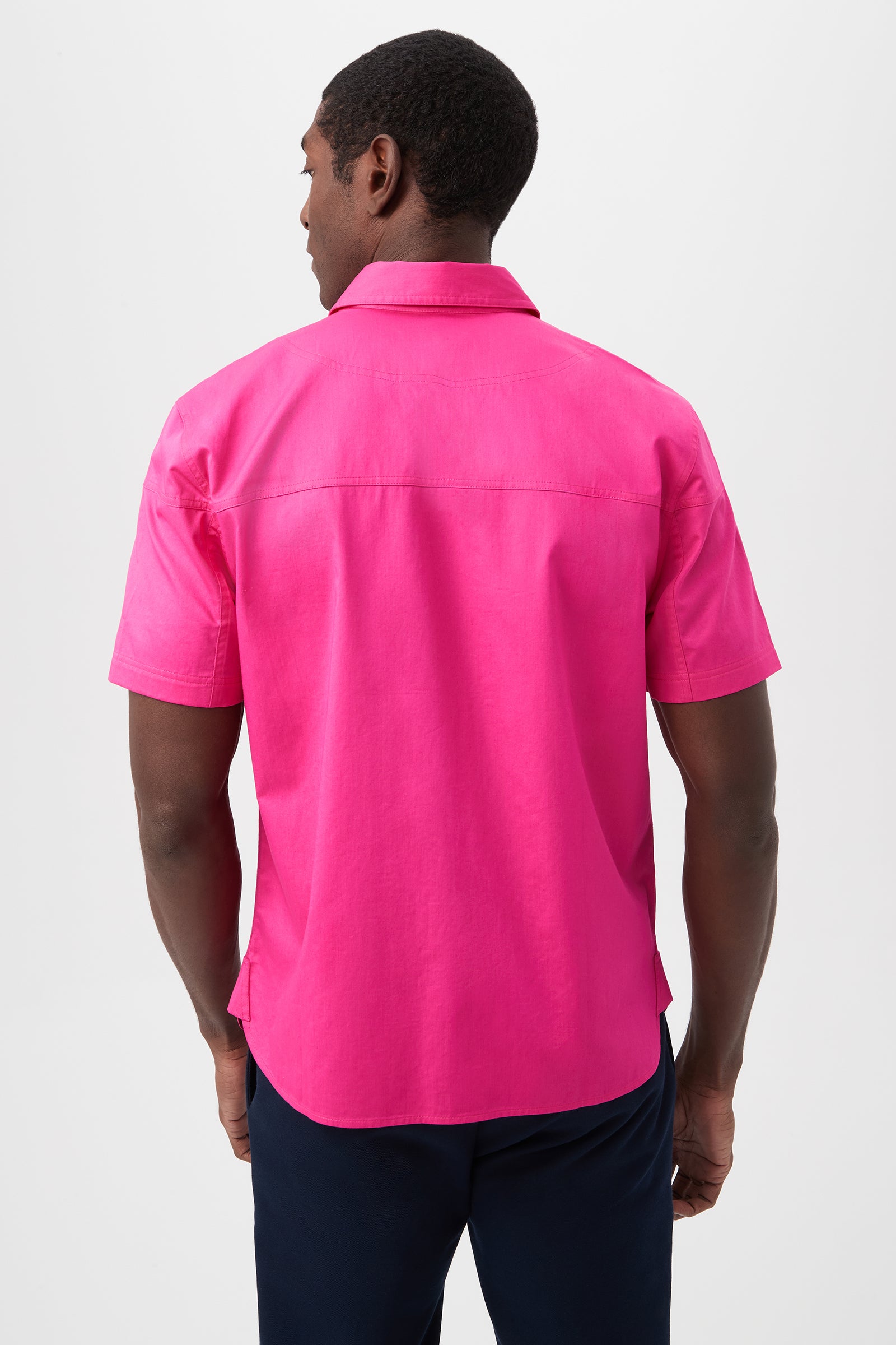 A man faces away, wearing the Shelton Short-Sleeve Shirt in bright pink cotton sateen with dark pants, standing against a plain white background.