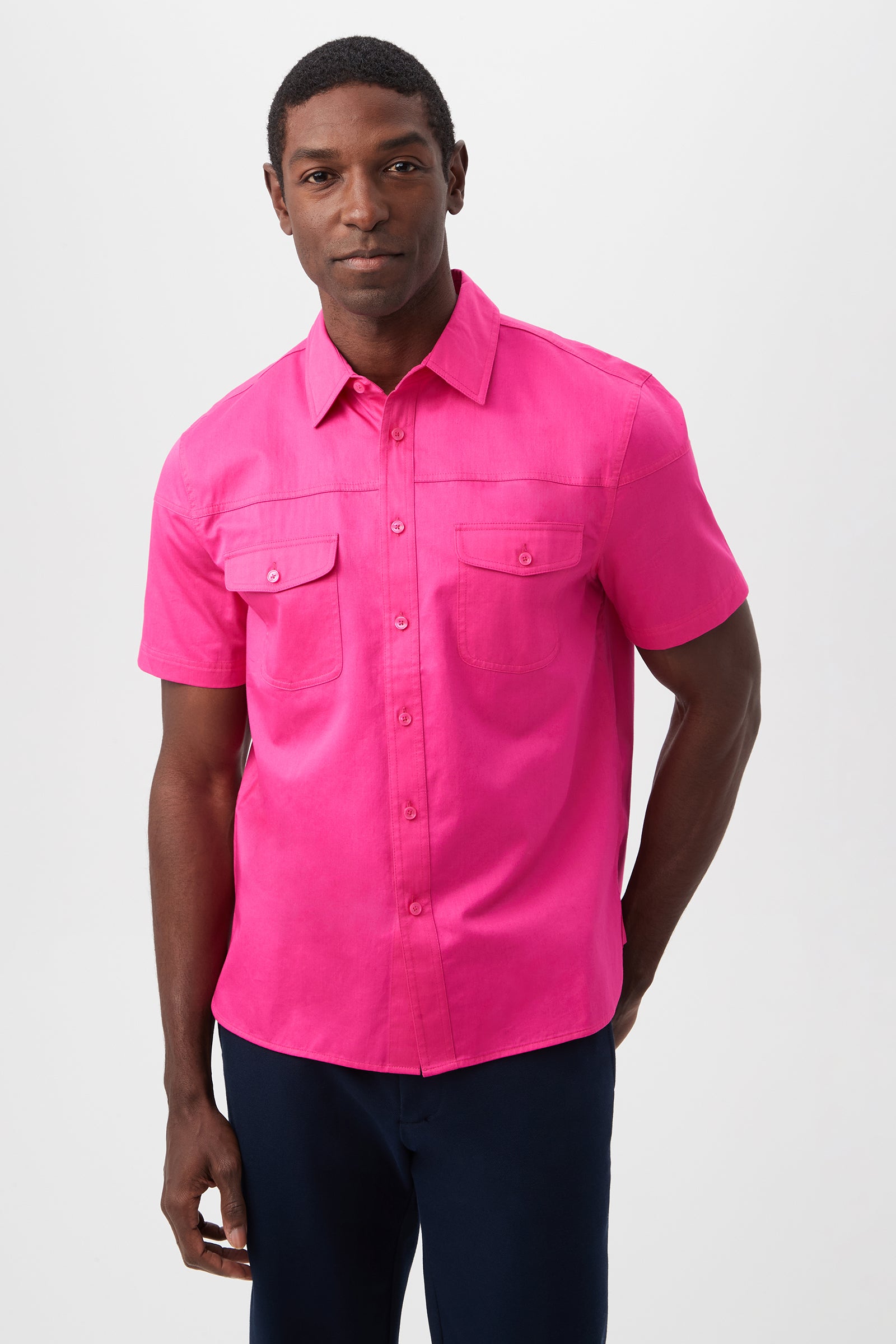 A man stands against a plain background wearing the Shelton Short-Sleeve Shirt in Petal Pink cotton sateen, featuring two front pockets, paired with dark pants.