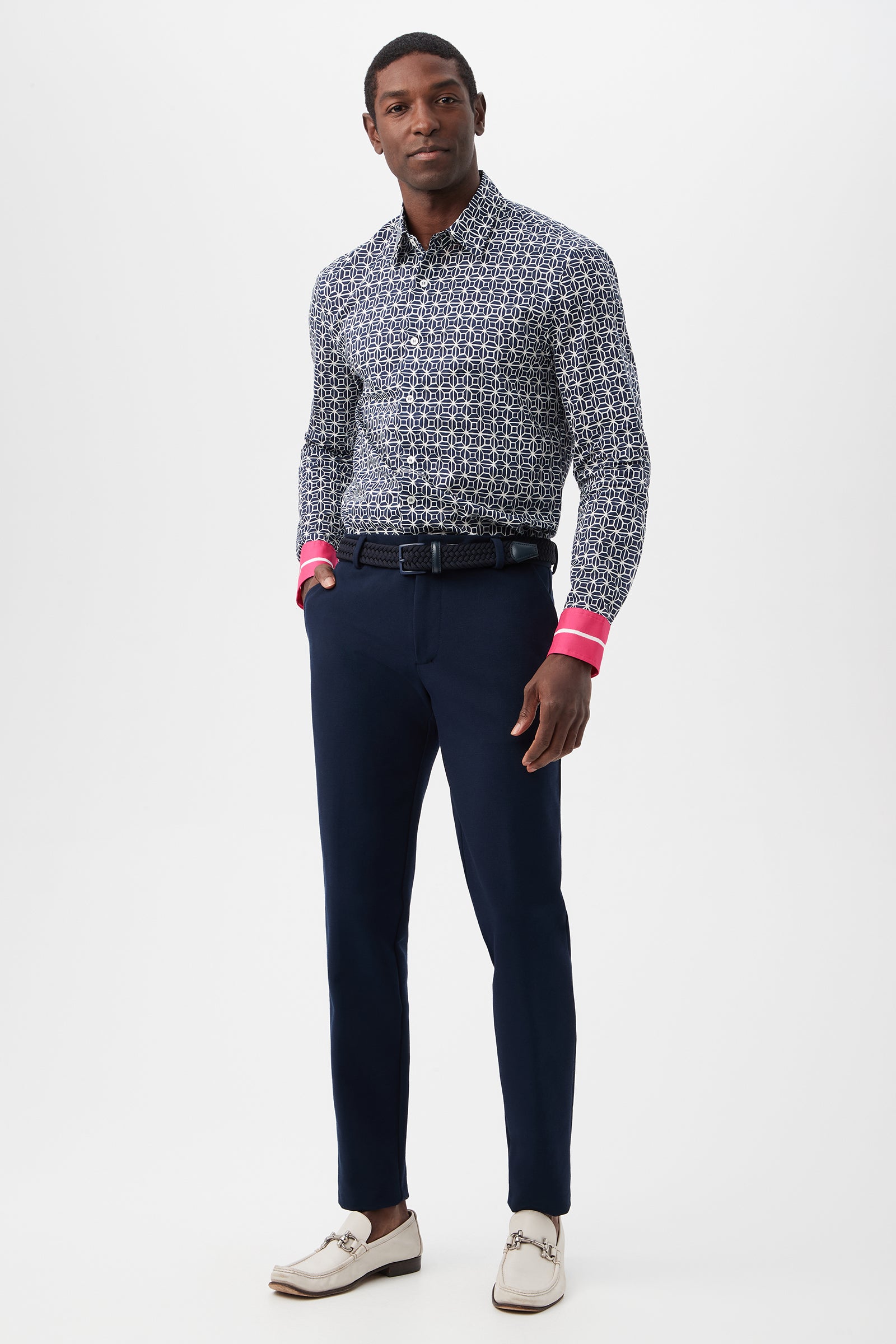 A man stands against a white background wearing a patterned button-up shirt with pink cuffs, the Clyde Slim Trouser - Stretch Piqué in navy blue, a belt, and white dress shoes.