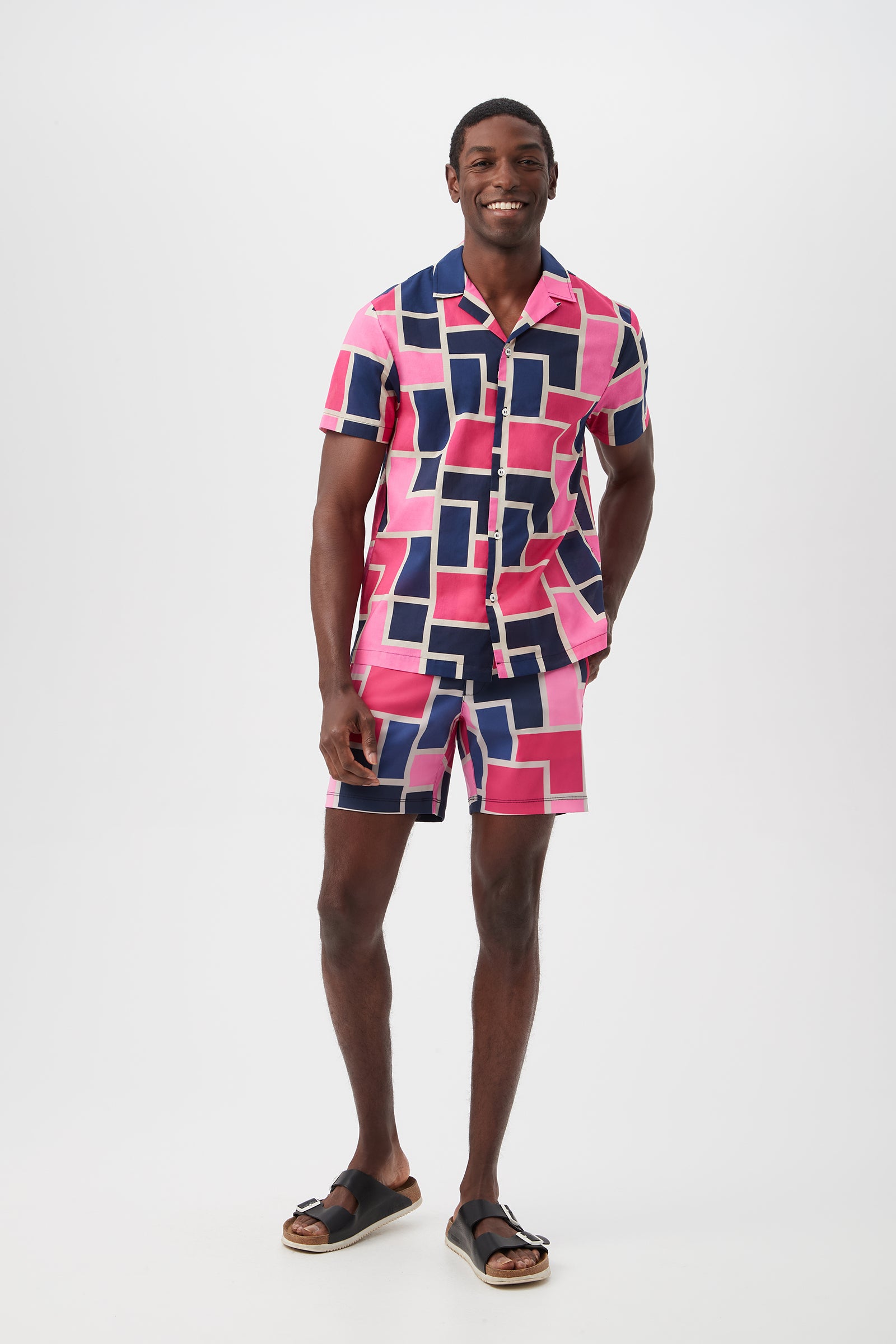 A man smiles wearing the Honolulu Board Short - Block Party, featuring a bold geometric pink, navy, and white pattern. He pairs it with a matching short-sleeve shirt and black sandals, standing against a plain white background.