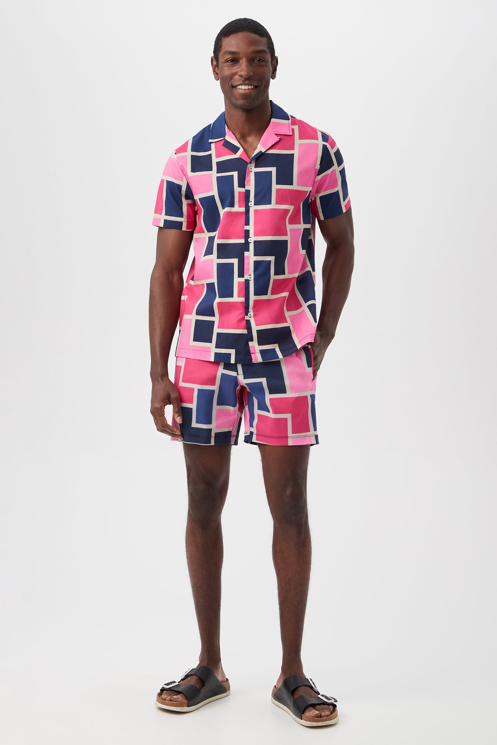 A man smiles wearing the Felix Camp Shirt - Block Party, a pink, navy, and white geometric short-sleeve shirt and matching shorts set with black sandals, standing against a plain white background.