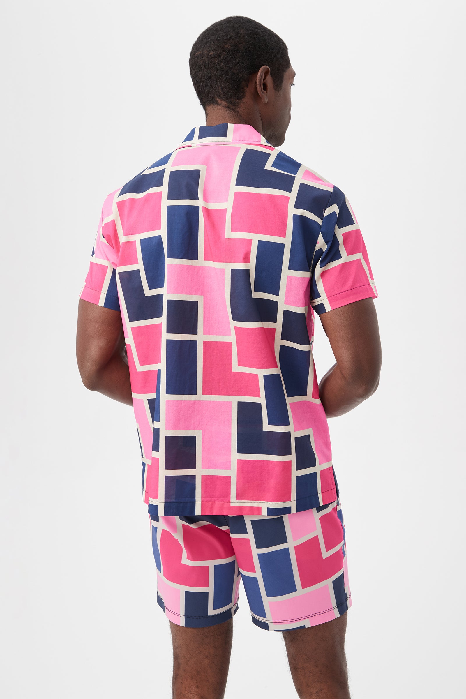 A man is seen from behind wearing the Felix Camp Shirt - Block Party, a bright short-sleeve set featuring a geometric pattern of pink, navy blue, and white rectangles on a white background.