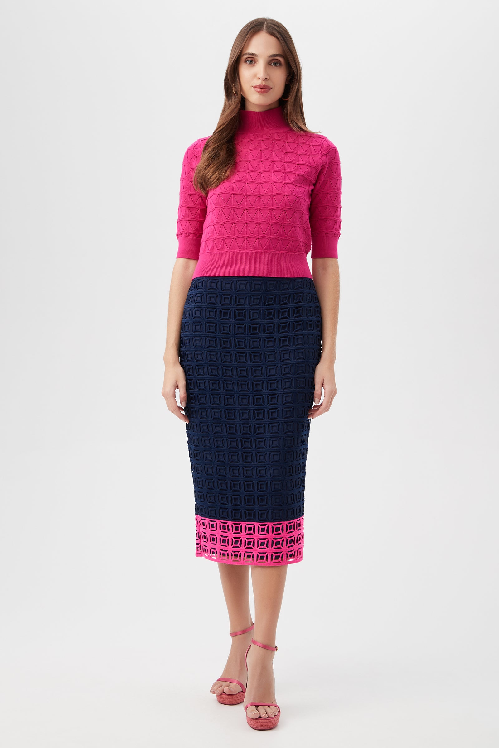 A woman with long hair wears pink heels and the Dita 2 Midi Pencil Skirt - Block Party Lace, a navy blue midi skirt featuring a textured geometric pattern and pink border, styled with a bright pink textured short-sleeve top against a plain background.