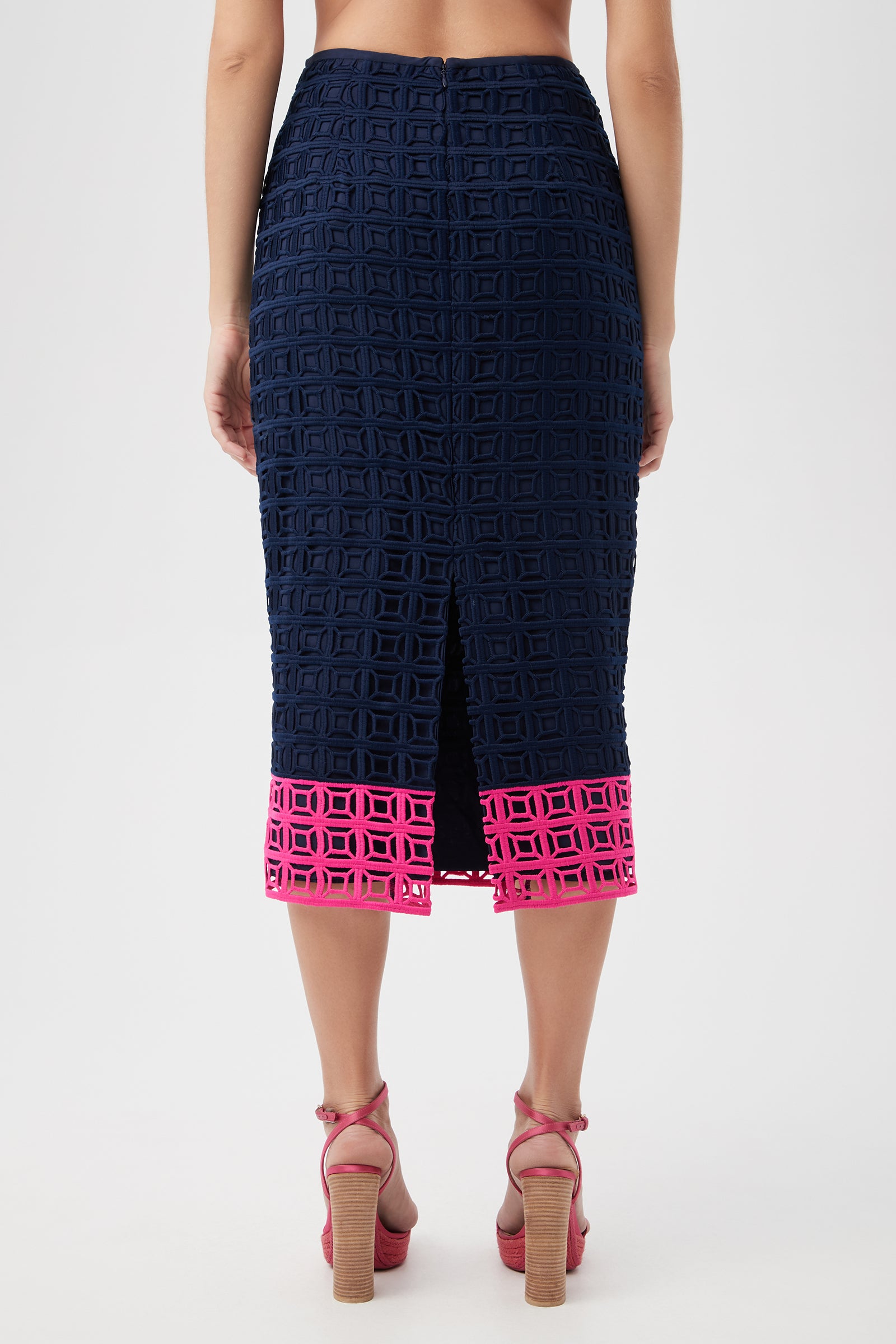 A woman is shown from the back wearing the Dita 2 Midi Pencil Skirt - Block Party Lace, featuring navy geometric cutout lace, a center slit, and a vivid pink cutout border at the hem. She pairs it with pink high-heeled sandals.
