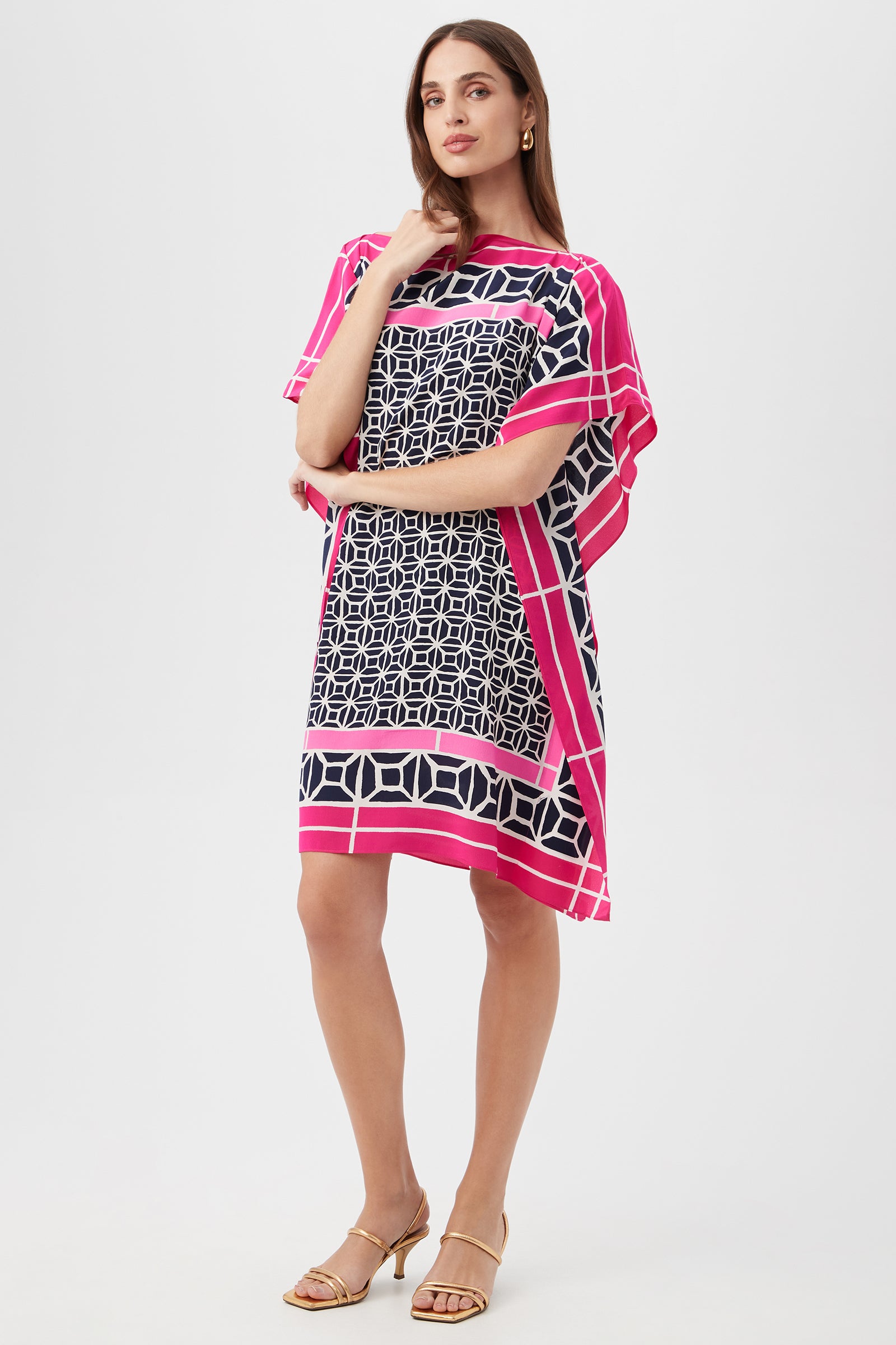 A woman models the Theodora Caftan Dress - Durell Silk in Multi, featuring a loose, knee-length silhouette with bold geometric patterns. She accessorizes with gold hoop earrings and tan heeled sandals against a plain white backdrop.