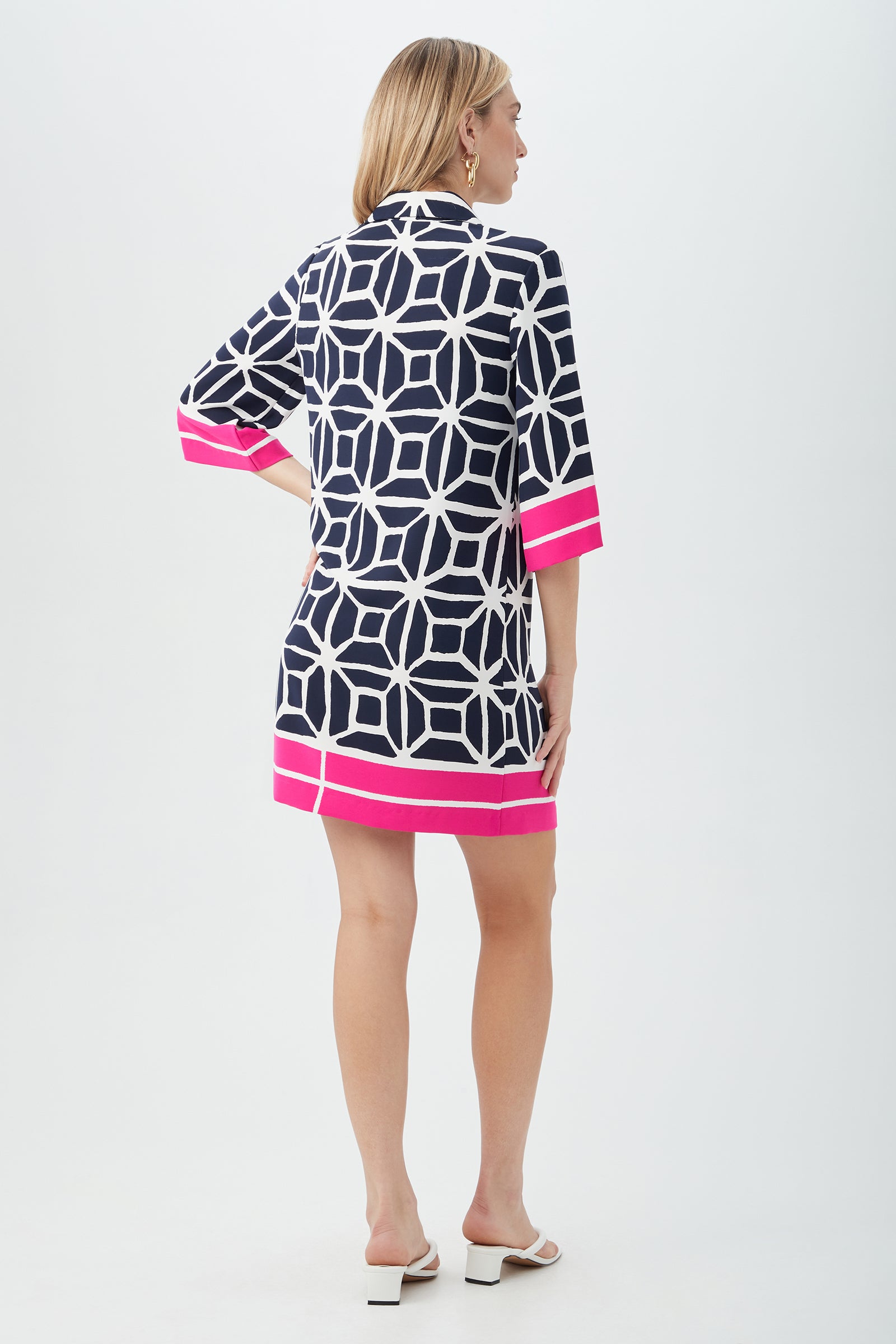 A woman with shoulder-length blonde hair faces away, wearing the Argina 3/4-Sleeve Dress in Durell Georgette—navy with a white geometric pattern and bright pink bands on the sleeves and hem—paired with white heeled sandals.