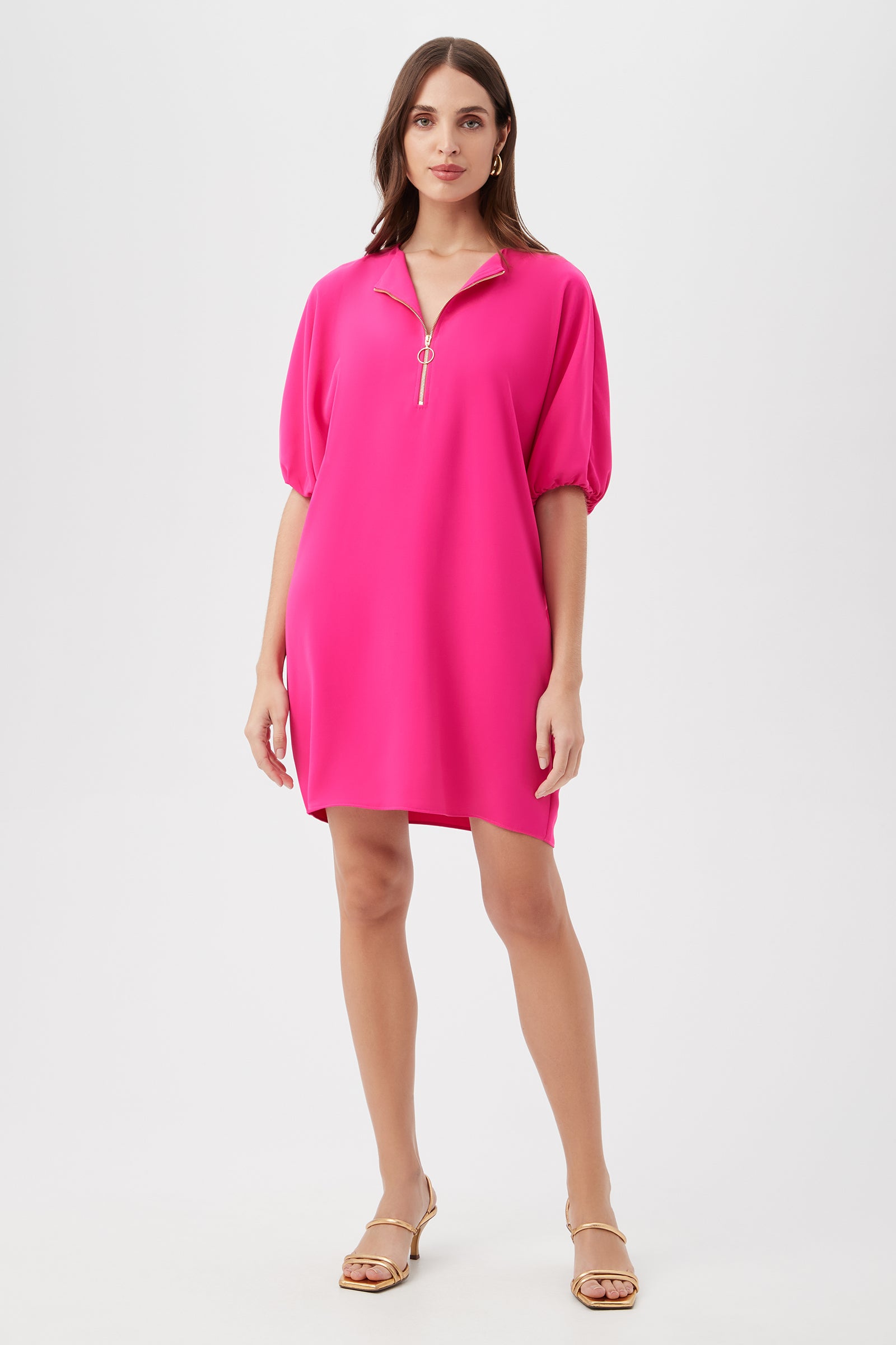 A woman with long brown hair models the Zariah Dress in Petal Pink, featuring a loose fit, elbow-length puff sleeves, and a front zipper, against a plain white background. She completes the look with flat strappy sandals.