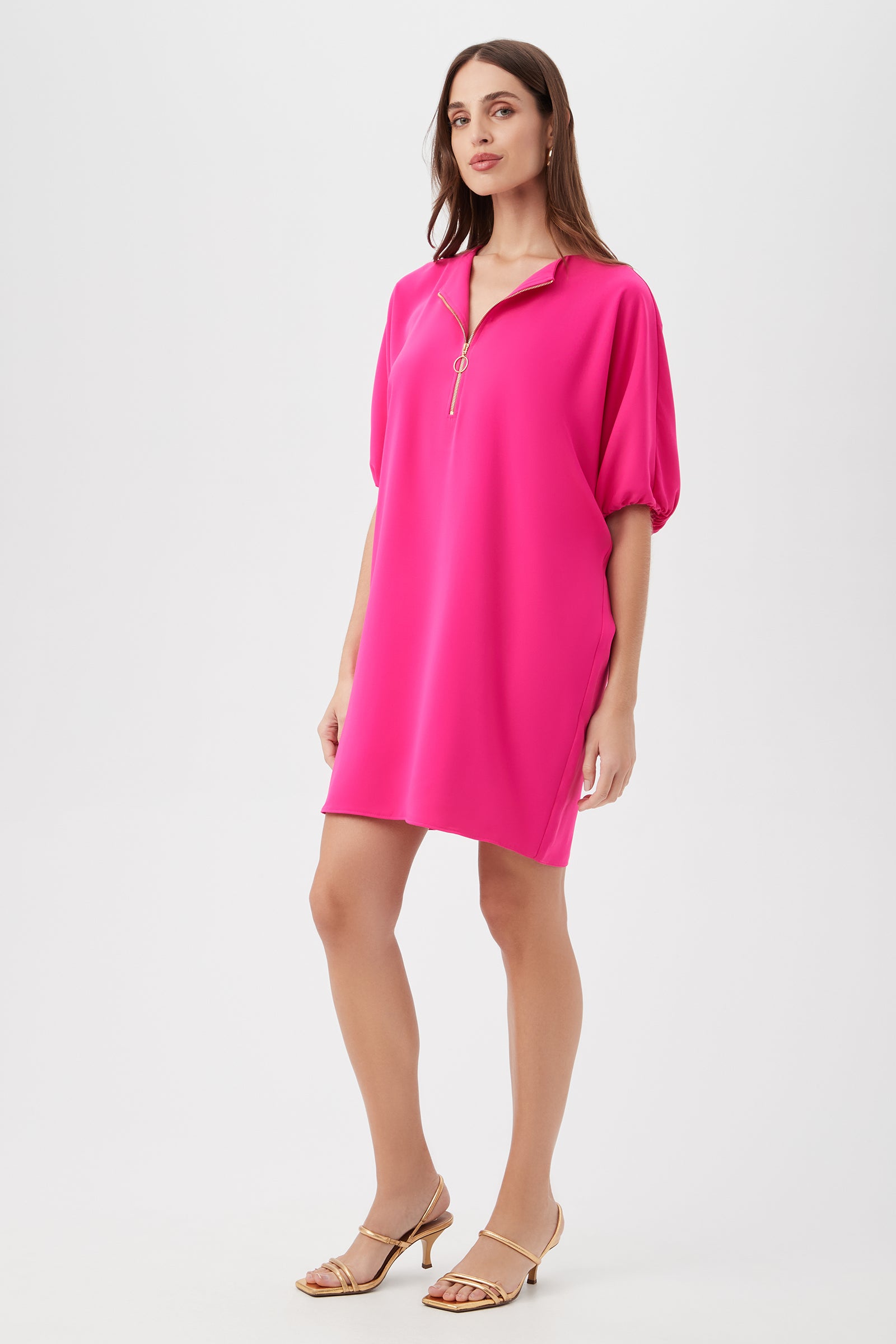 A woman with straight brown hair and neutral sandals stands against a white background wearing the Zariah Dress, a loose, bright pink short-sleeve style with a zip collar, gazing slightly to the side.