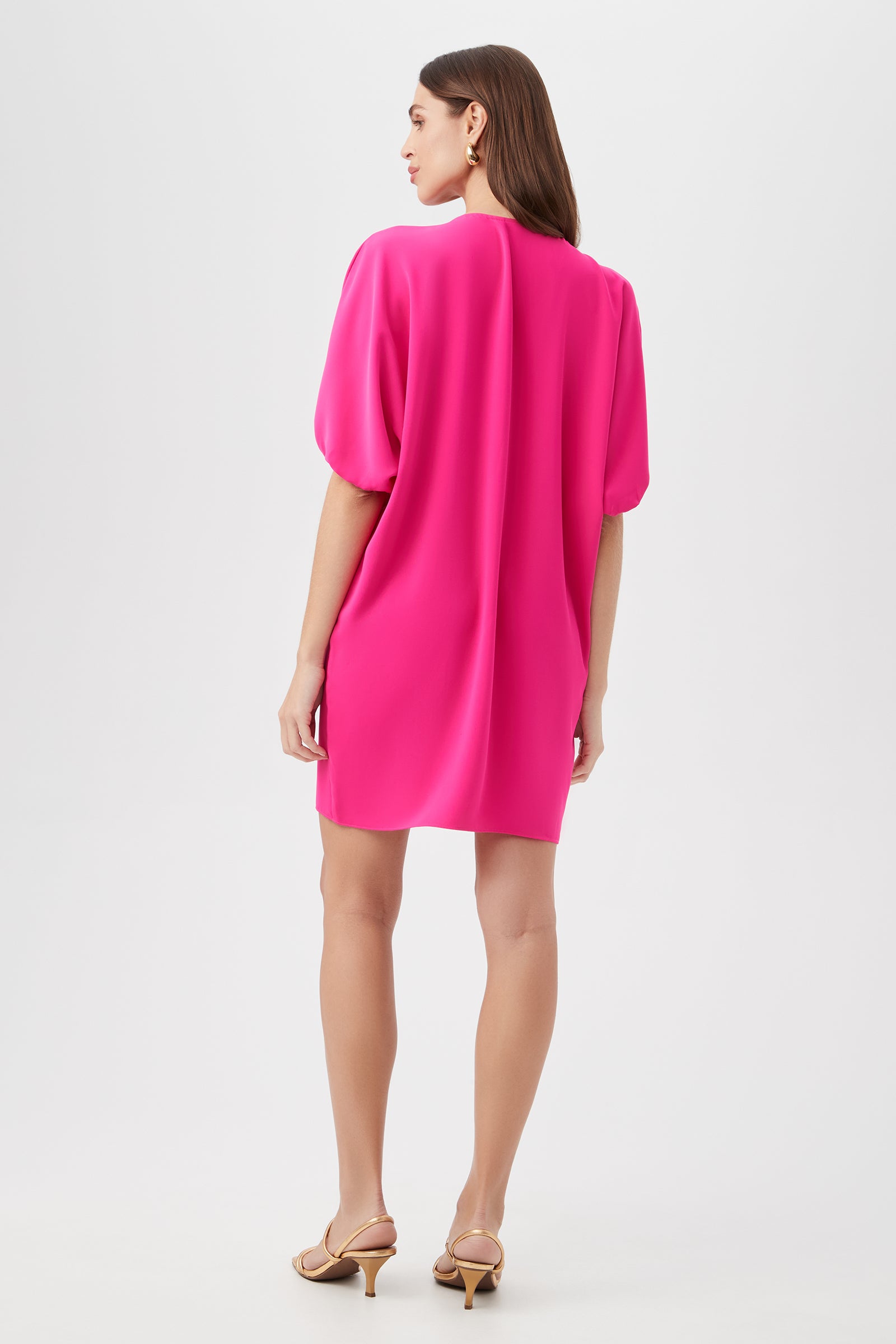 A woman faces away, modeling the Zariah Dress—a bright pink, loose-fit short dress with elbow-length sleeves and beige heeled sandals—set against a plain white background.