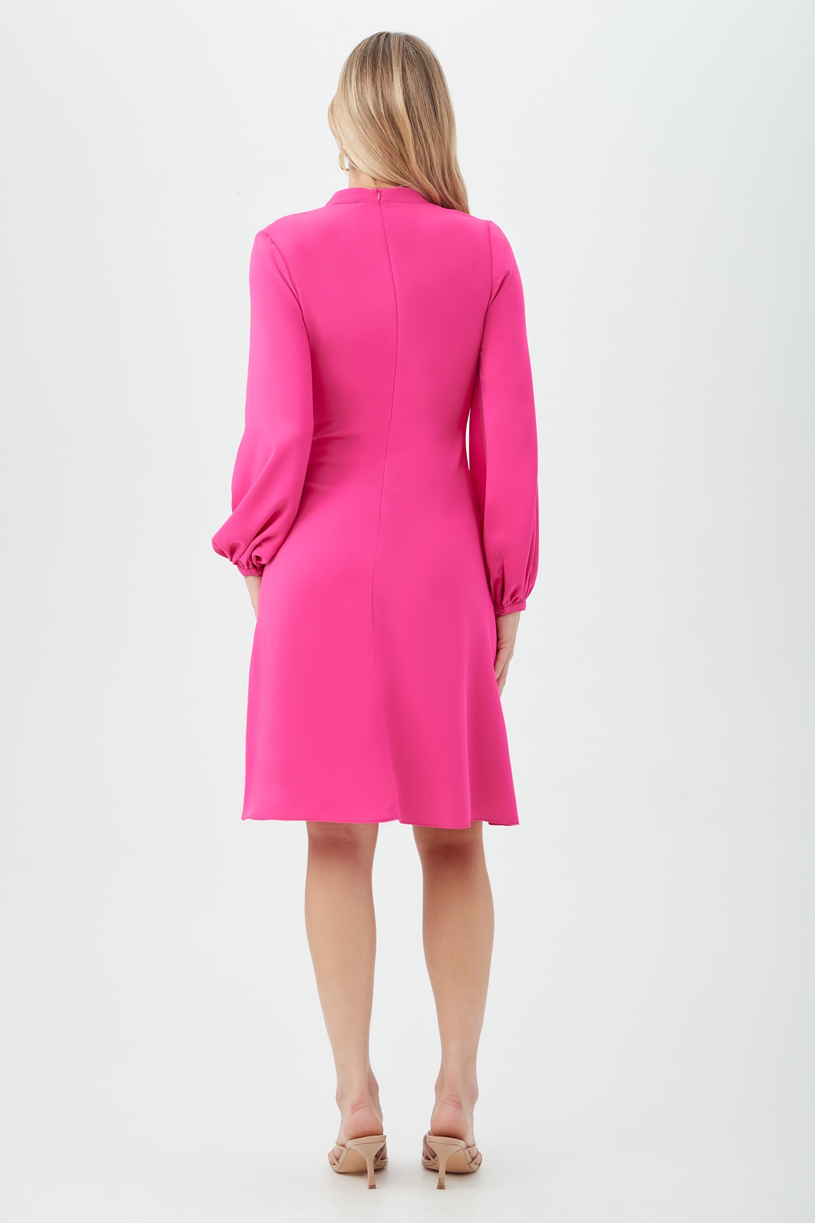 A woman with light hair faces away, wearing the Lora Dress—a knee-length, long-sleeved, bright pink dress with a relaxed fit and puffed sleeves—paired with nude heeled sandals against a plain white background.