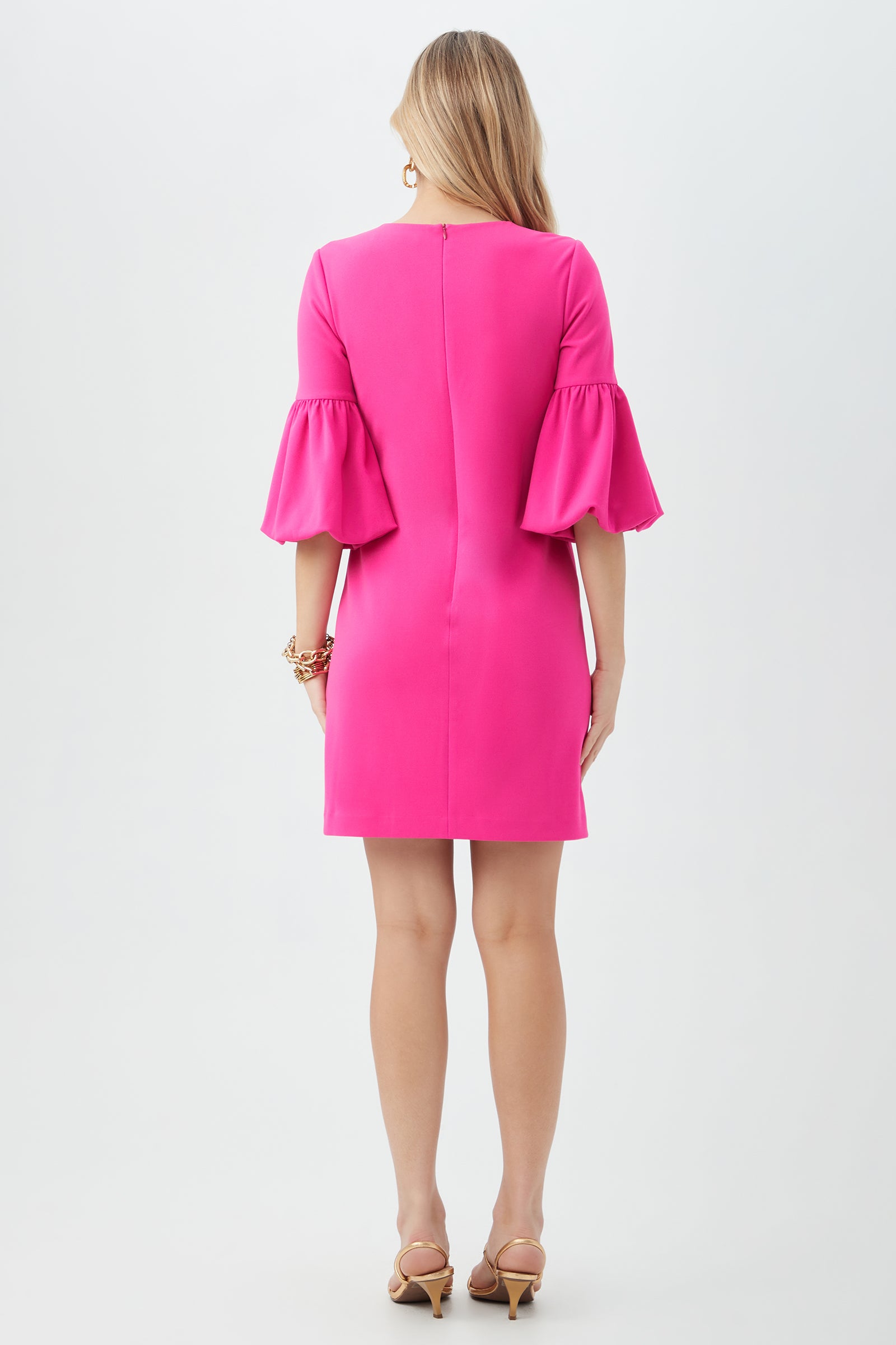 A woman with long blonde hair stands facing away in the Redford Dress, a bright pink style with ruffled bell sleeves. She pairs it with nude heels and bracelets, set against a plain white background.