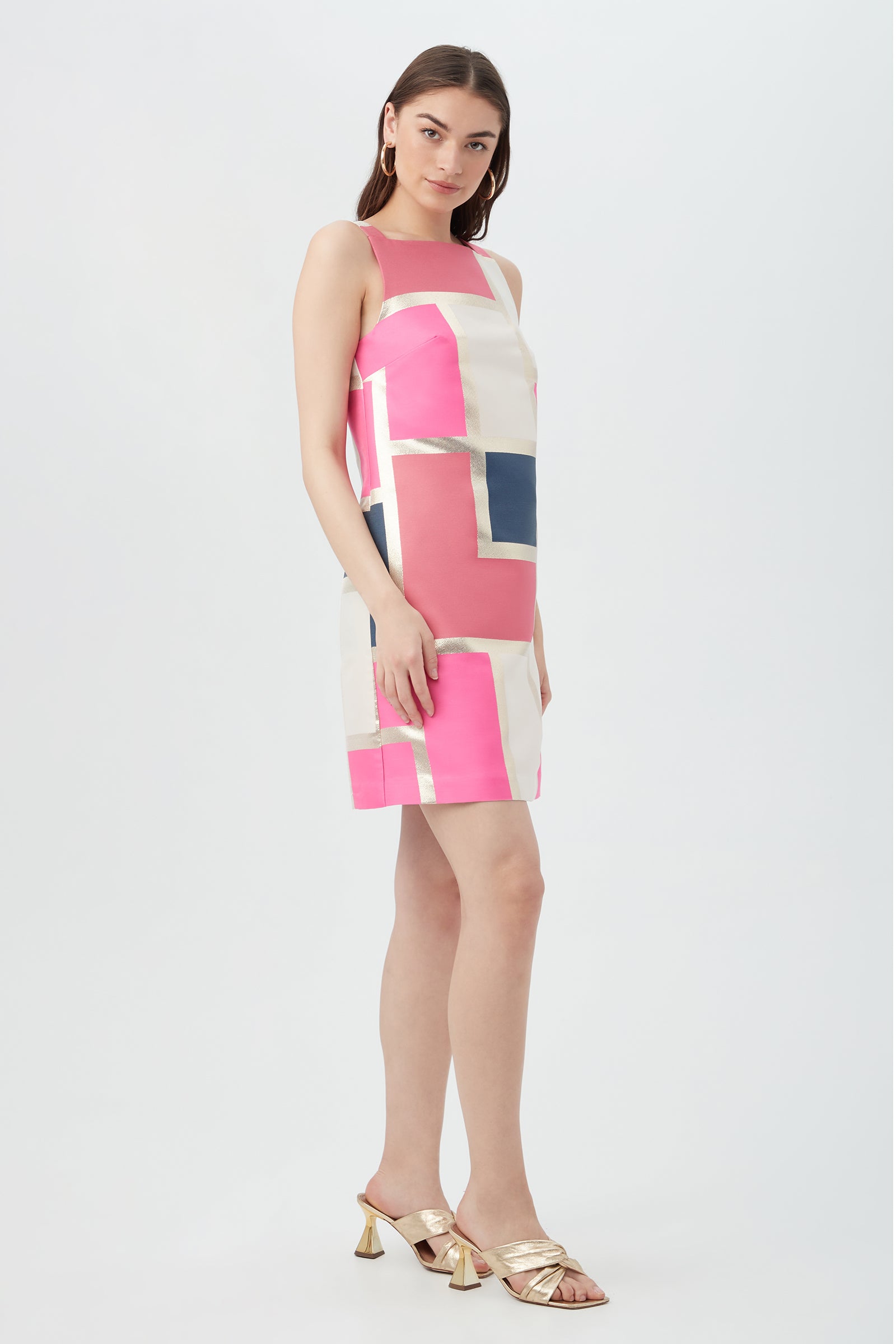 Woman wearing the Creosote Dress - Block Party Jacquard, a sleeveless, knee-length dress with bold pink, cream, navy, and metallic gold geometric patterns, paired with gold heeled sandals against a white background.