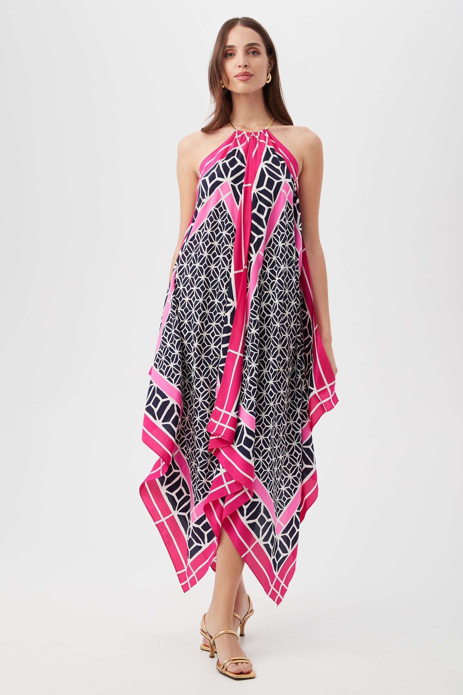 A woman stands confidently in heeled sandals against a white background, wearing the Justinian Midi Dress - Durell Silk in Multi, a flowing sleeveless dress with a geometric black and white pattern and a bold pink border.