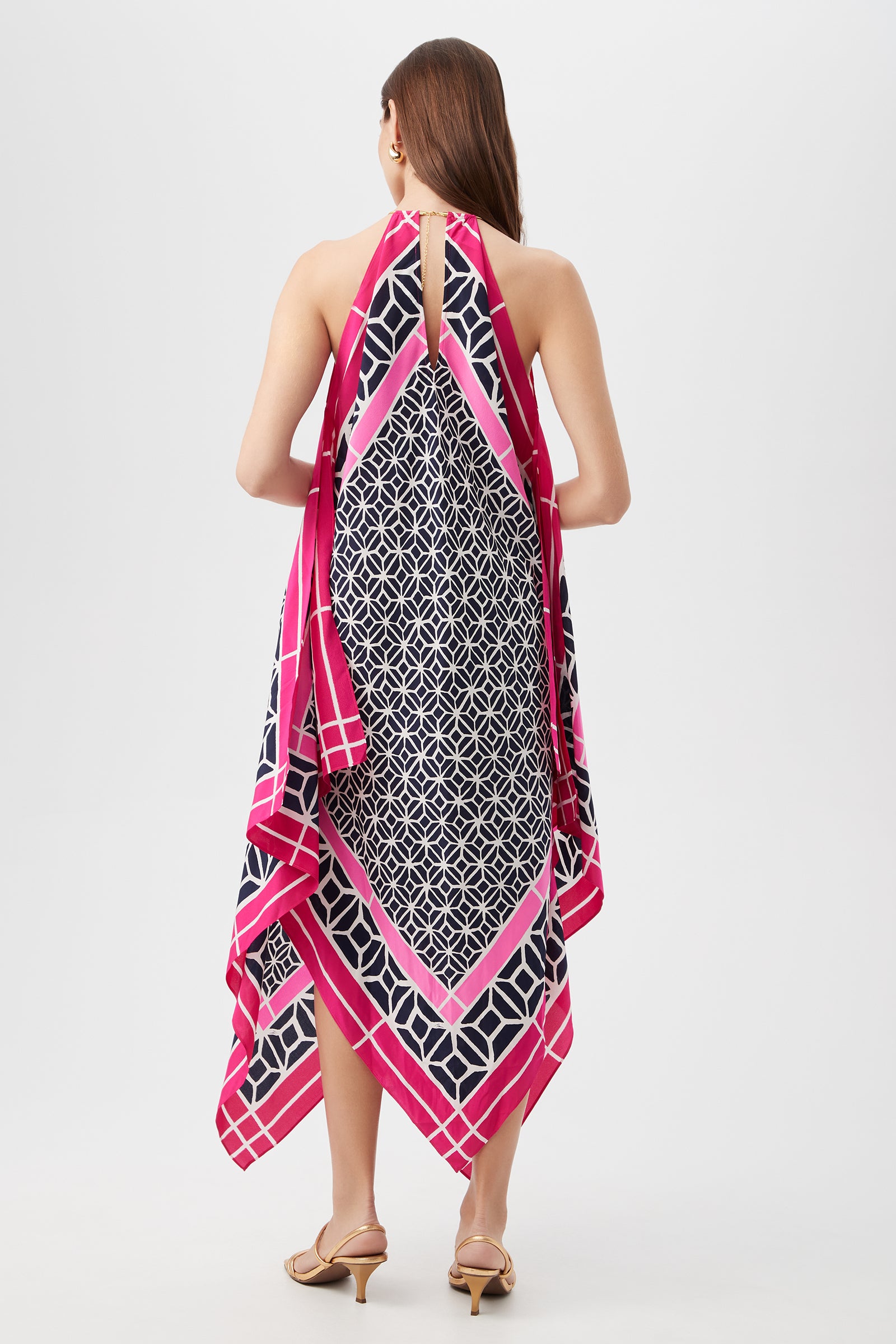 A woman with long brown hair faces away from the camera, wearing the Justinian Midi Dress - Durell Silk, a halter-neck dress featuring a geometric navy and white print with pink and white borders. She pairs it with beige heeled sandals.