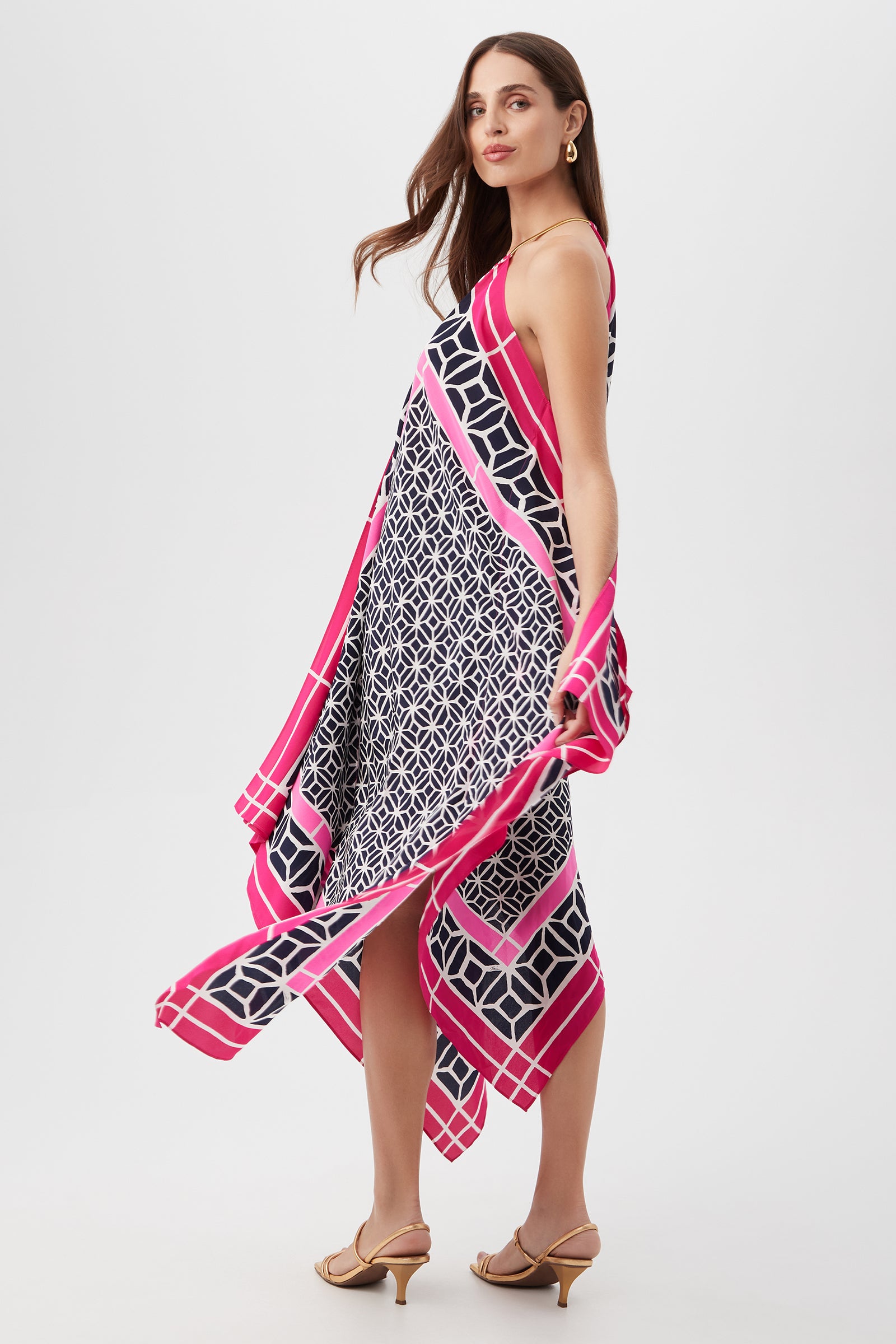 A woman in tan heeled sandals models the Justinian Midi Dress - Durell Silk, a loose, sleeveless handkerchief dress featuring a navy and white geometric pattern with bold pink and white striped borders, against a plain light background.