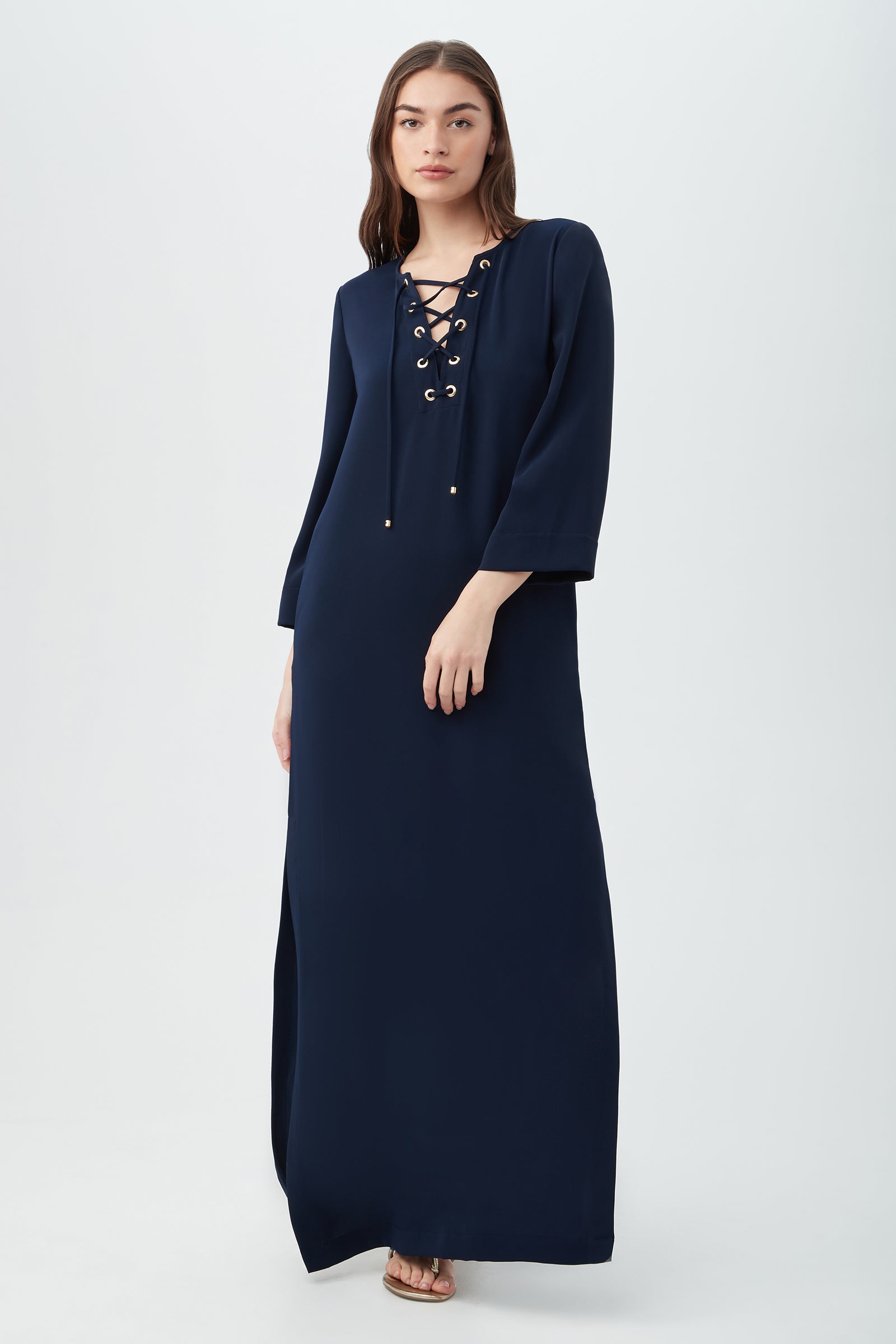A woman poses against a plain light background wearing the Catamaran 3/4-Sleeve Maxi Dress in indigo, featuring a lace-up front neckline. She has straight brown hair and sandals.