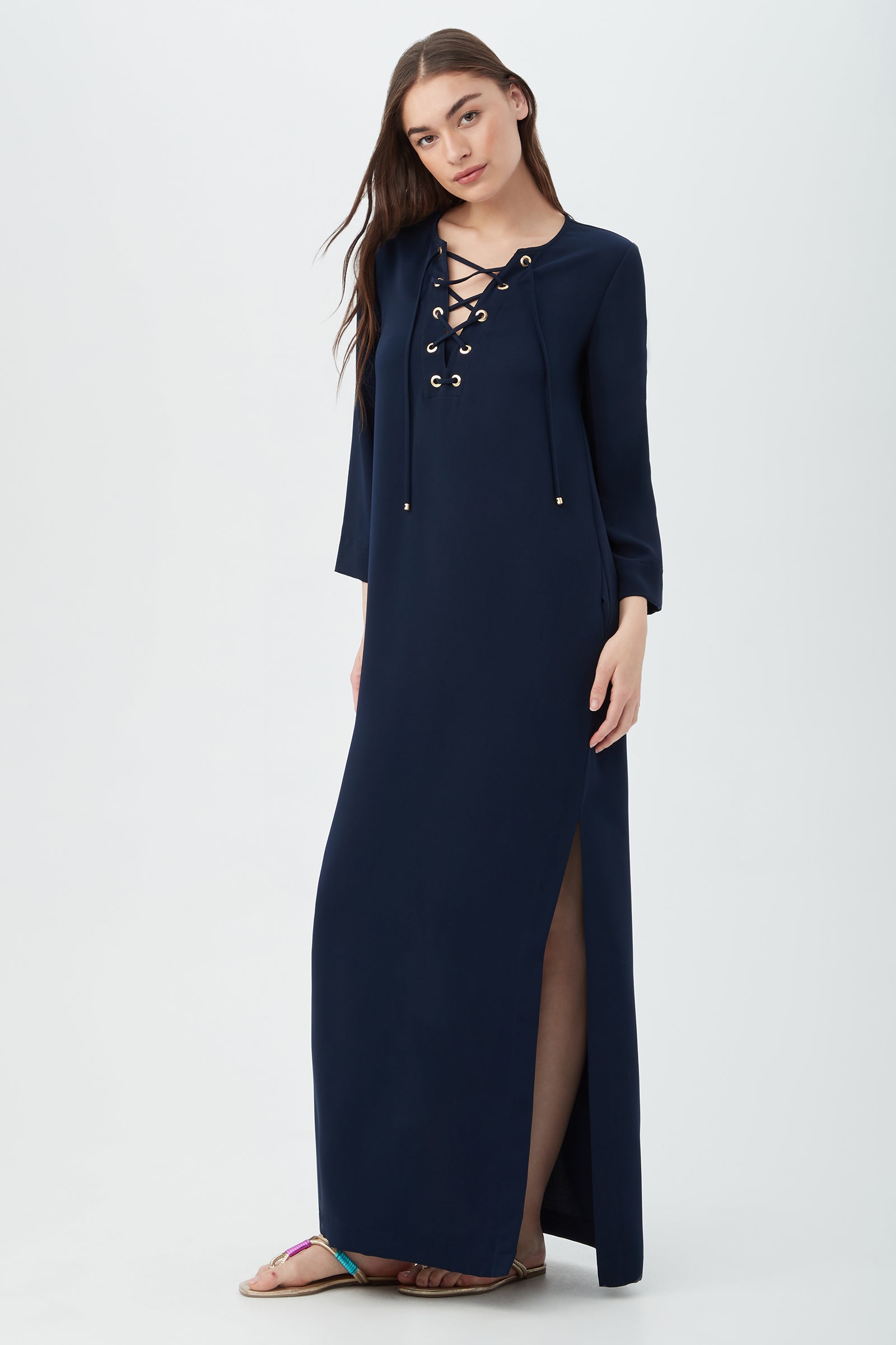 A woman with long, straight brown hair wears the Catamaran 3/4-Sleeve Maxi Dress in navy blue, featuring a lace-up neckline and thigh-high slit, paired with flat sandals, standing against a plain light background.