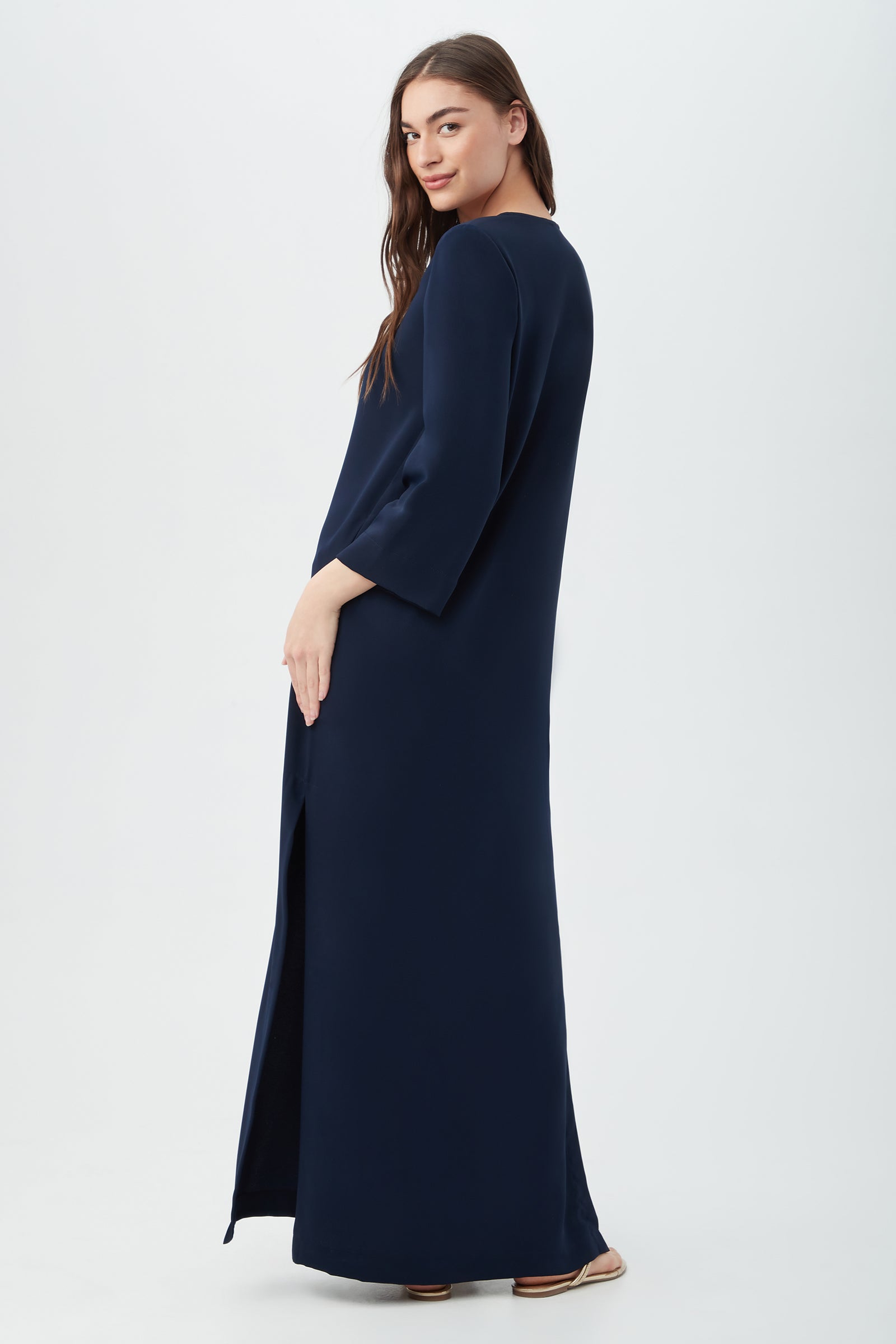 A woman with long brown hair stands sideways, smiling over her shoulder as she models the Catamaran 3/4-Sleeve Maxi Dress in navy blue with a side slit against a plain white background.