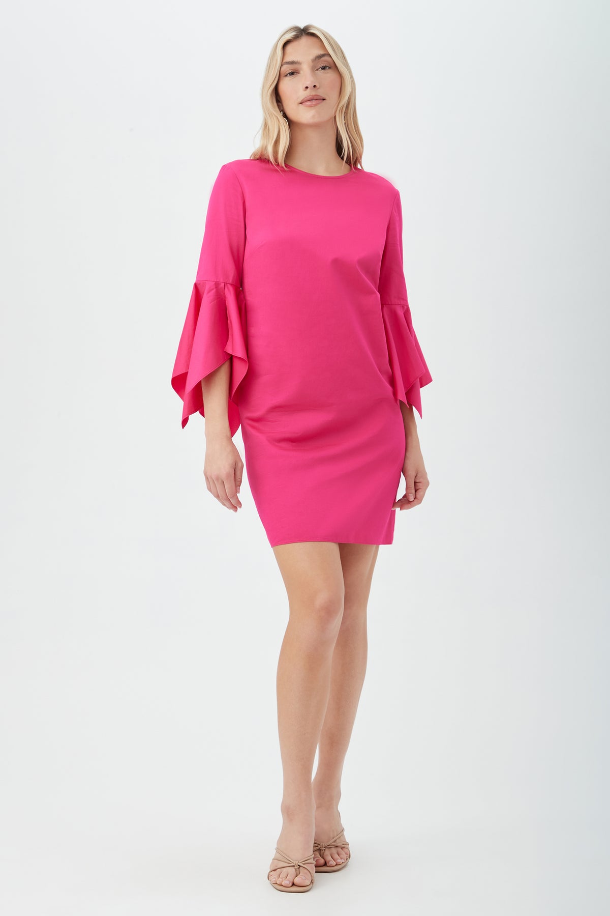 A woman with long, light hair and a relaxed, confident posture wears the Fontainebleau Dress—a bright pink, knee-length dress with ruffled bell sleeves—paired with beige sandals against a plain background.