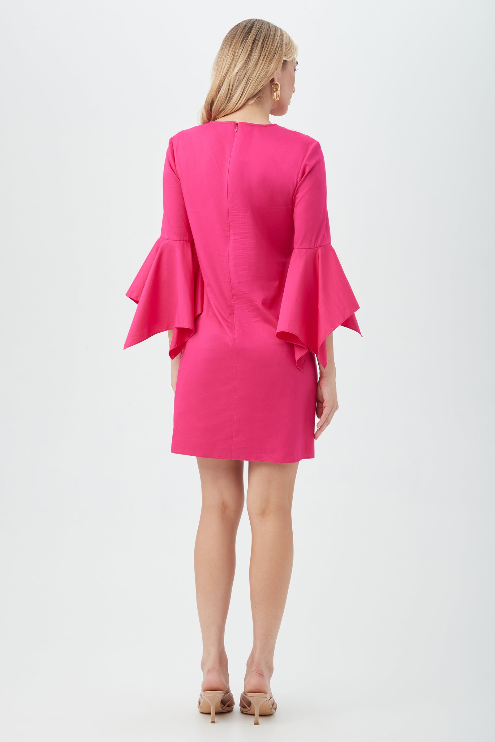 A woman with long blonde hair faces away, wearing the Fontainebleau Dress—a bright pink, knee-length style with flared ruffled sleeves—paired with nude open-toe heels, set against a plain white background.