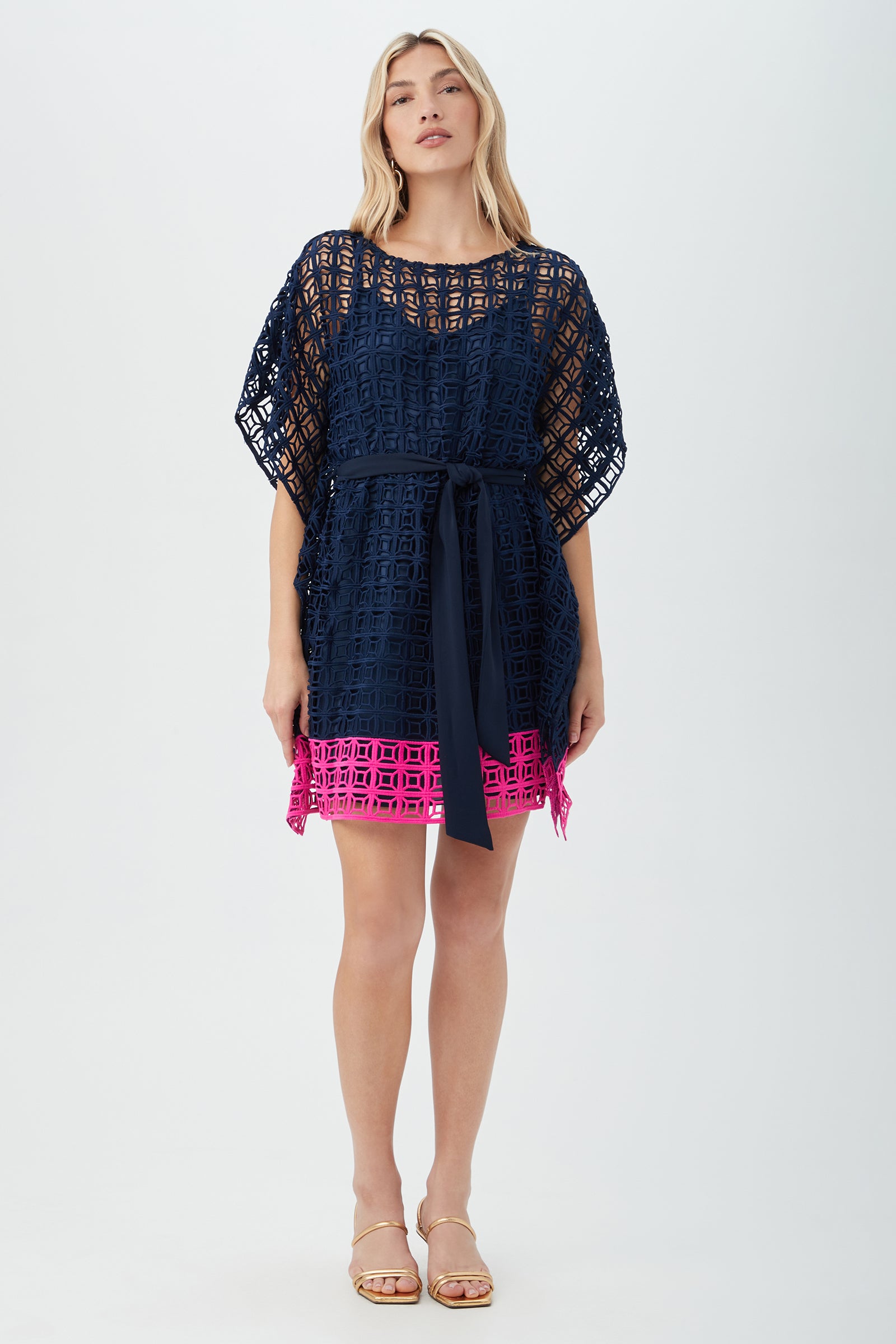 A woman with blonde hair stands facing forward against a white background, wearing the Dora Caftan Dress - Block Party Lace in indigo/petal pink, featuring geometric lace and a bright pink hem. She completes her look with sandals.