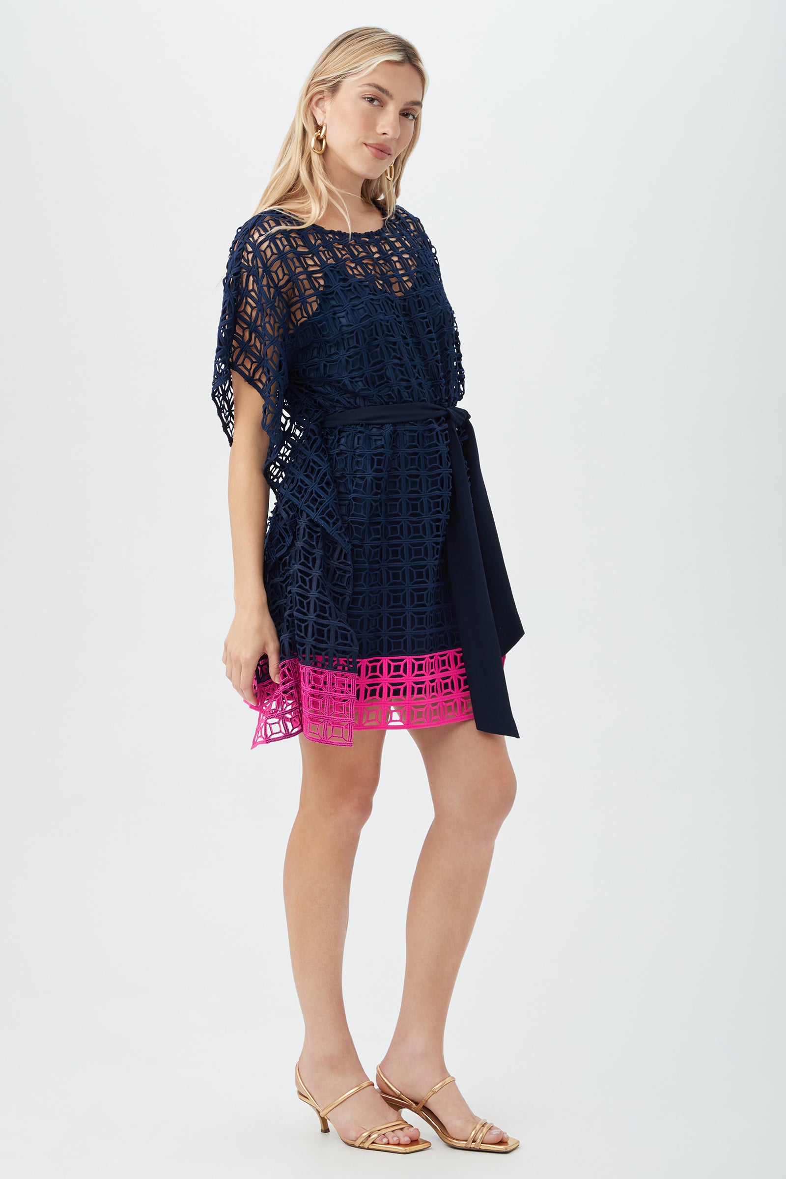 A woman wears the Dora Caftan Dress - Block Party Lace, a short-sleeved navy dress with geometric cutouts and a pink hem, belted at the waist. She accessorizes with hoop earrings, gold sandals, and has long blonde hair.