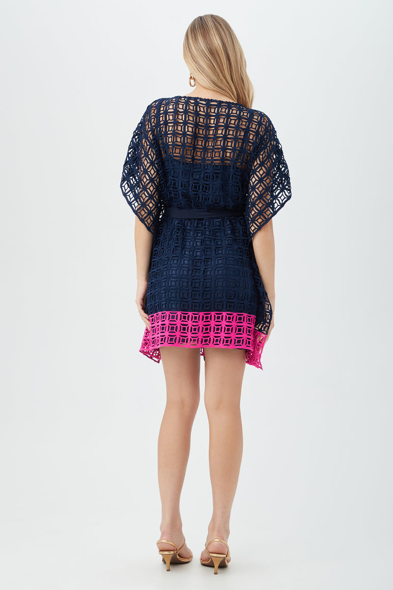 A woman with long blonde hair faces away, wearing the Dora Caftan Dress - Block Party Lace featuring a short, navy blue and pink geometric pattern with kimono sleeves and nude heels, set against a plain light background.