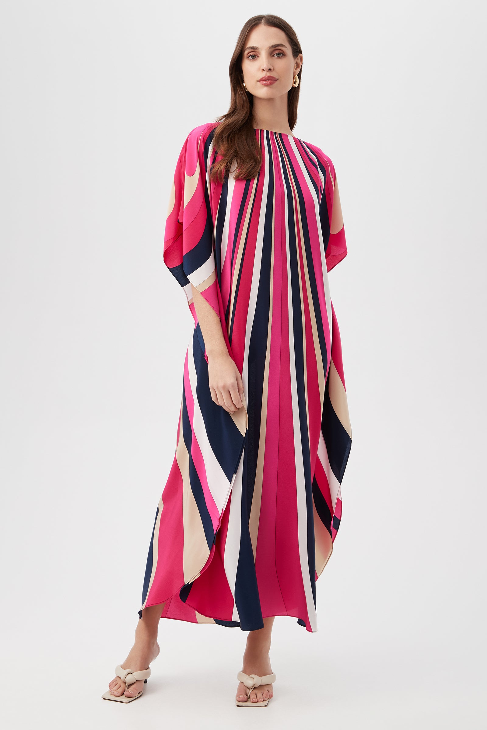A woman with long brown hair stands confidently against a white background wearing the Jalani Maxi Caftan Dress - Wynwood Waves Silk in Petal Pink Multi, a loose, floor-length style with bold vertical stripes and open-toe sandals.
