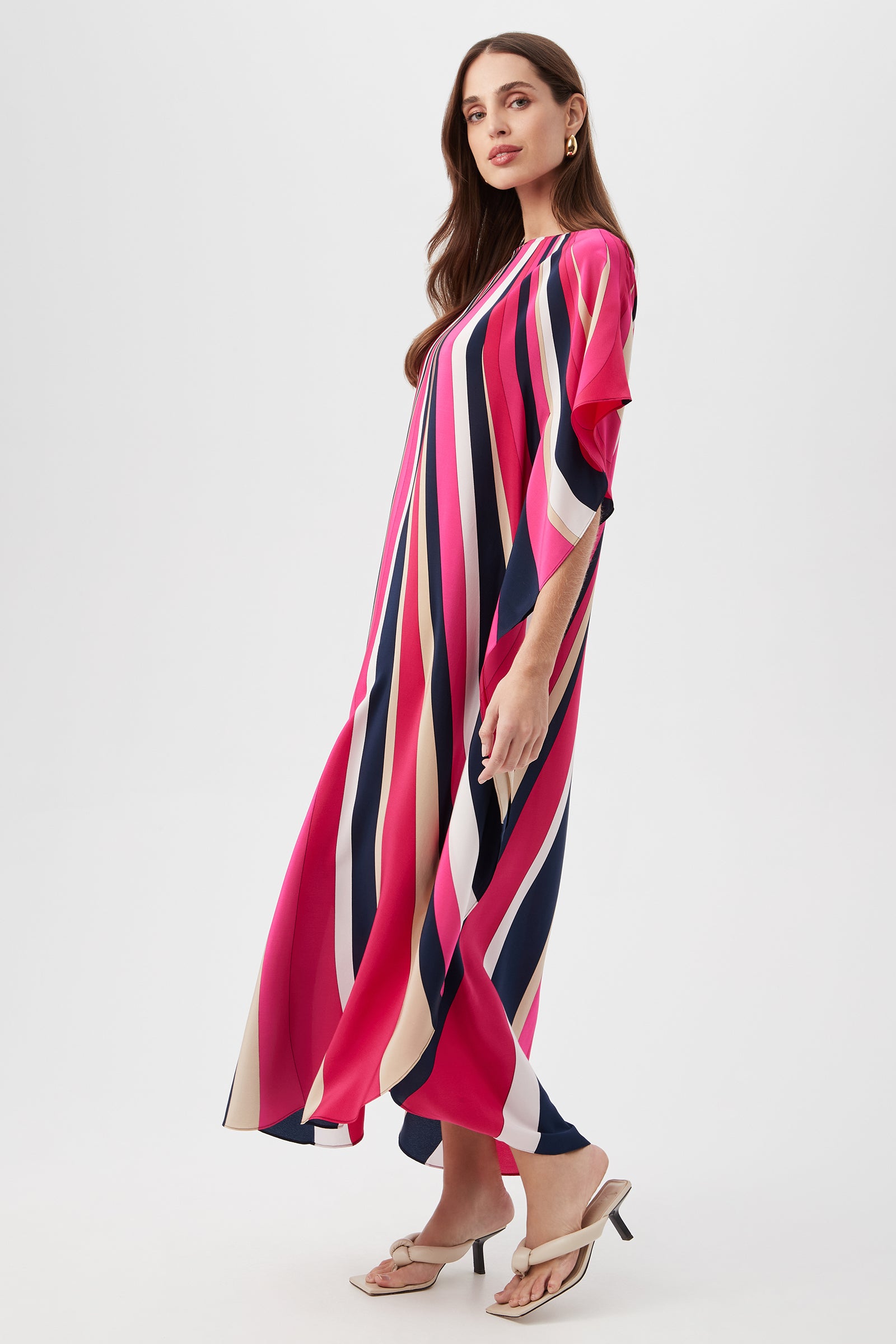 Woman wearing the Jalani Maxi Caftan Dress - Wynwood Waves Silk, a long flowing caftan with bold vertical stripes in pink, navy, cream, and coral, standing sideways with long brown hair and neutral sandals against a plain background.