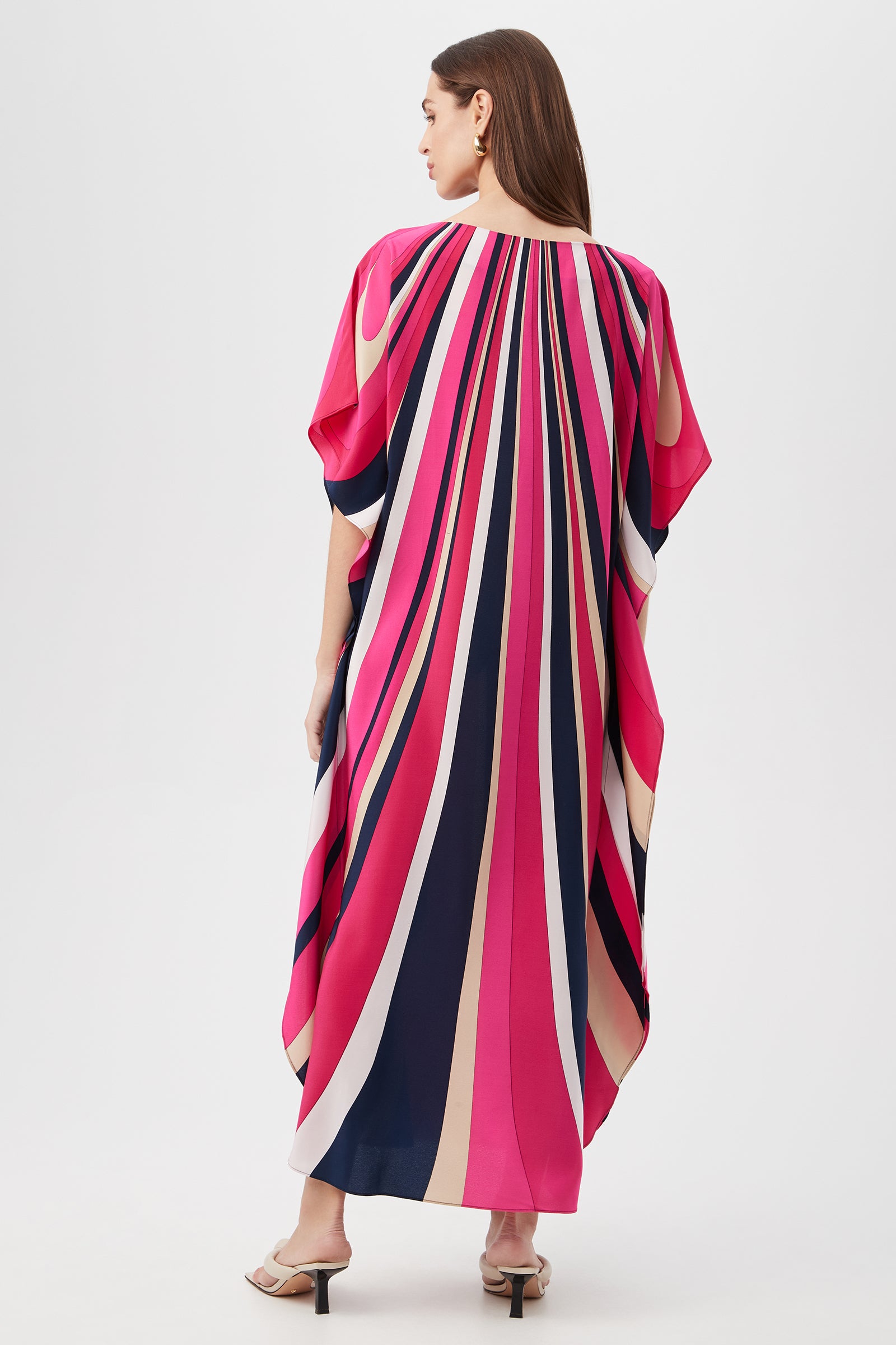 A woman with straight brown hair faces away, modeling the Jalani Maxi Caftan Dress - Wynwood Waves Silk, a loose-fitting, ankle-length kaftan with bold pink, navy, beige, and red vertical stripes. She wears open-toed heels against a plain white background.