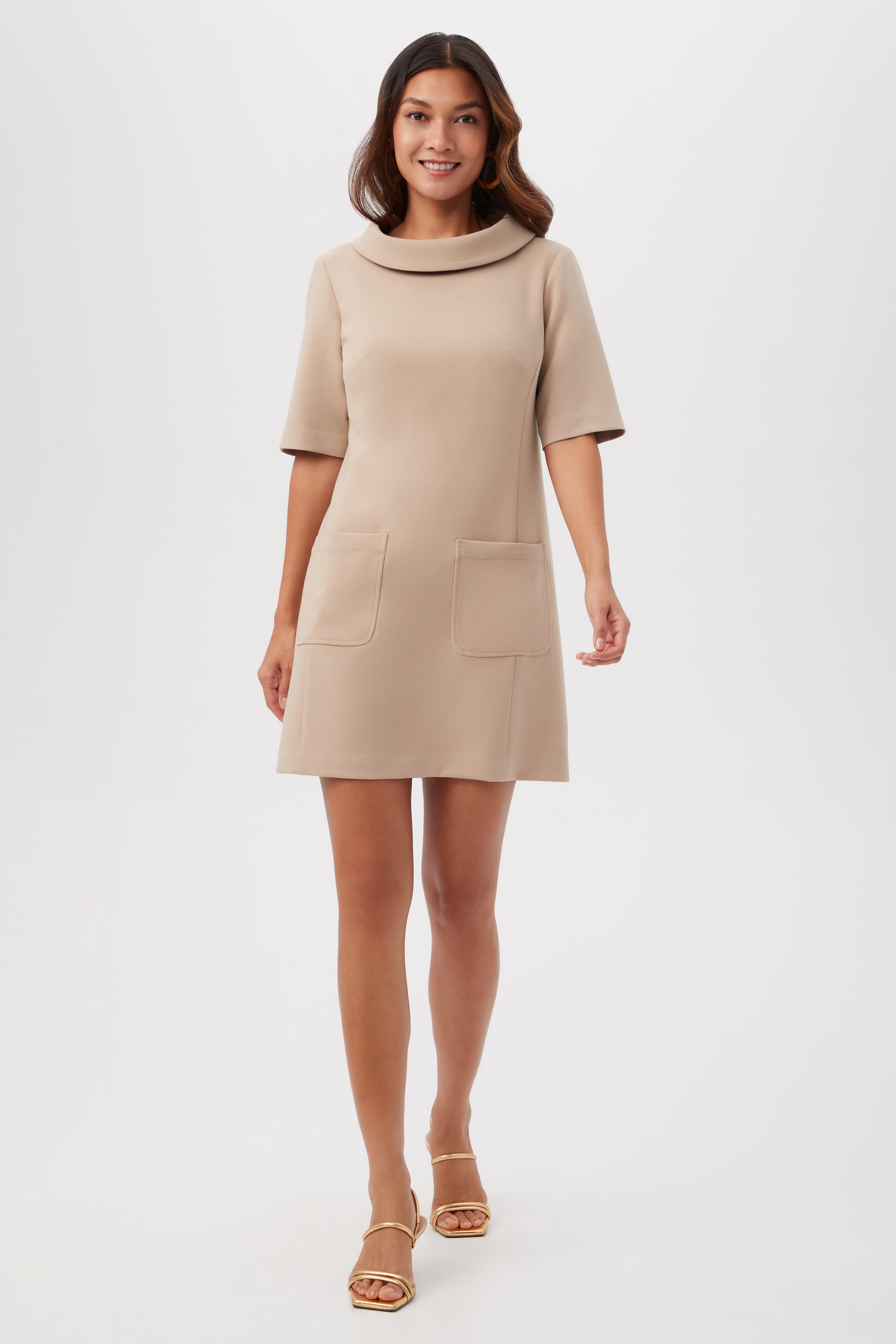 A woman stands and smiles against a white background wearing the Maleko Dress, a beige, knee-length design with short sleeves and two front pockets, paired with open-toe sandals.