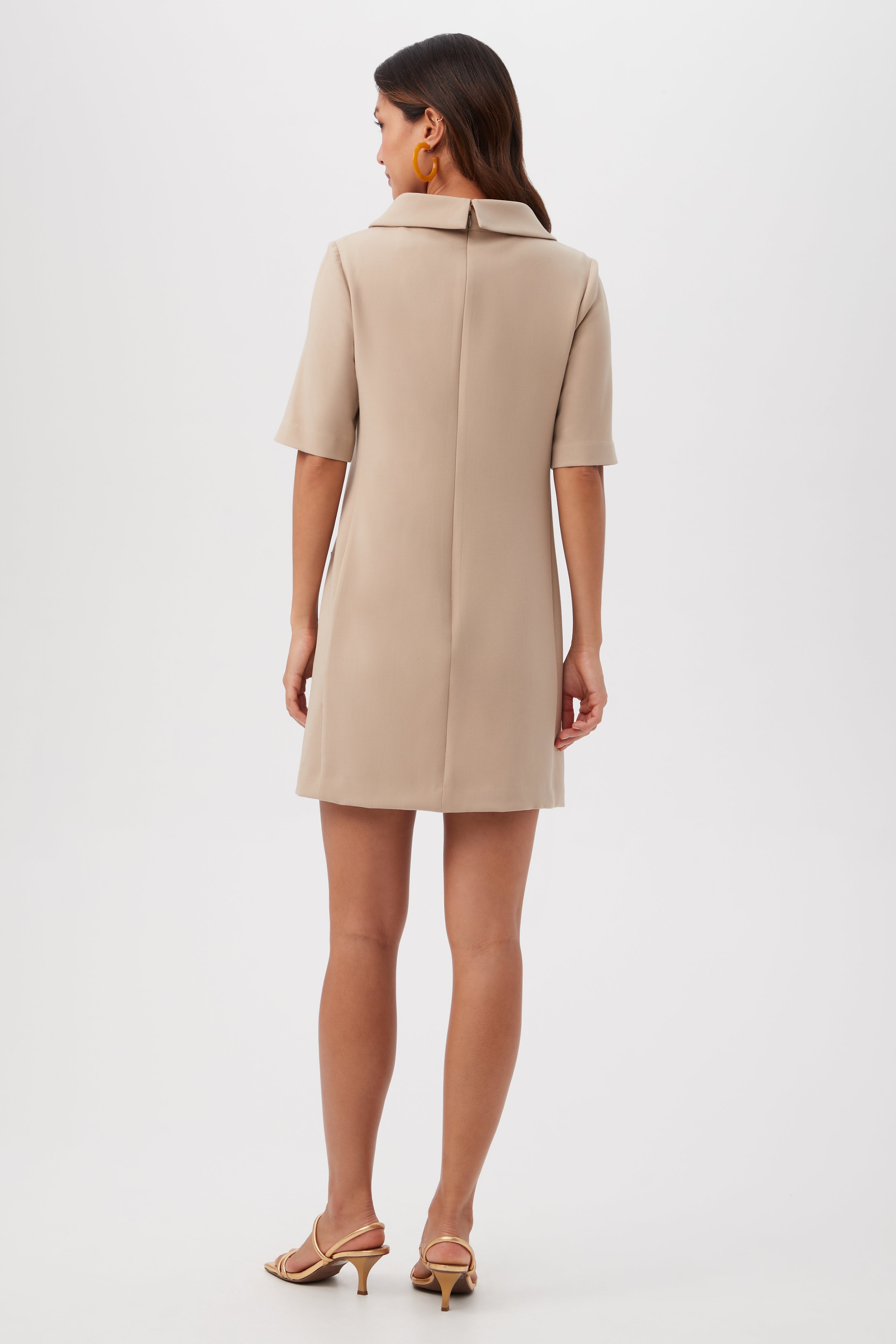 A woman faces away, wearing the beige, short-sleeved Maleko Dress with a collar and beige heeled sandals. Her hair is loose, she's accessorized with large hoop earrings, and the background is plain white.